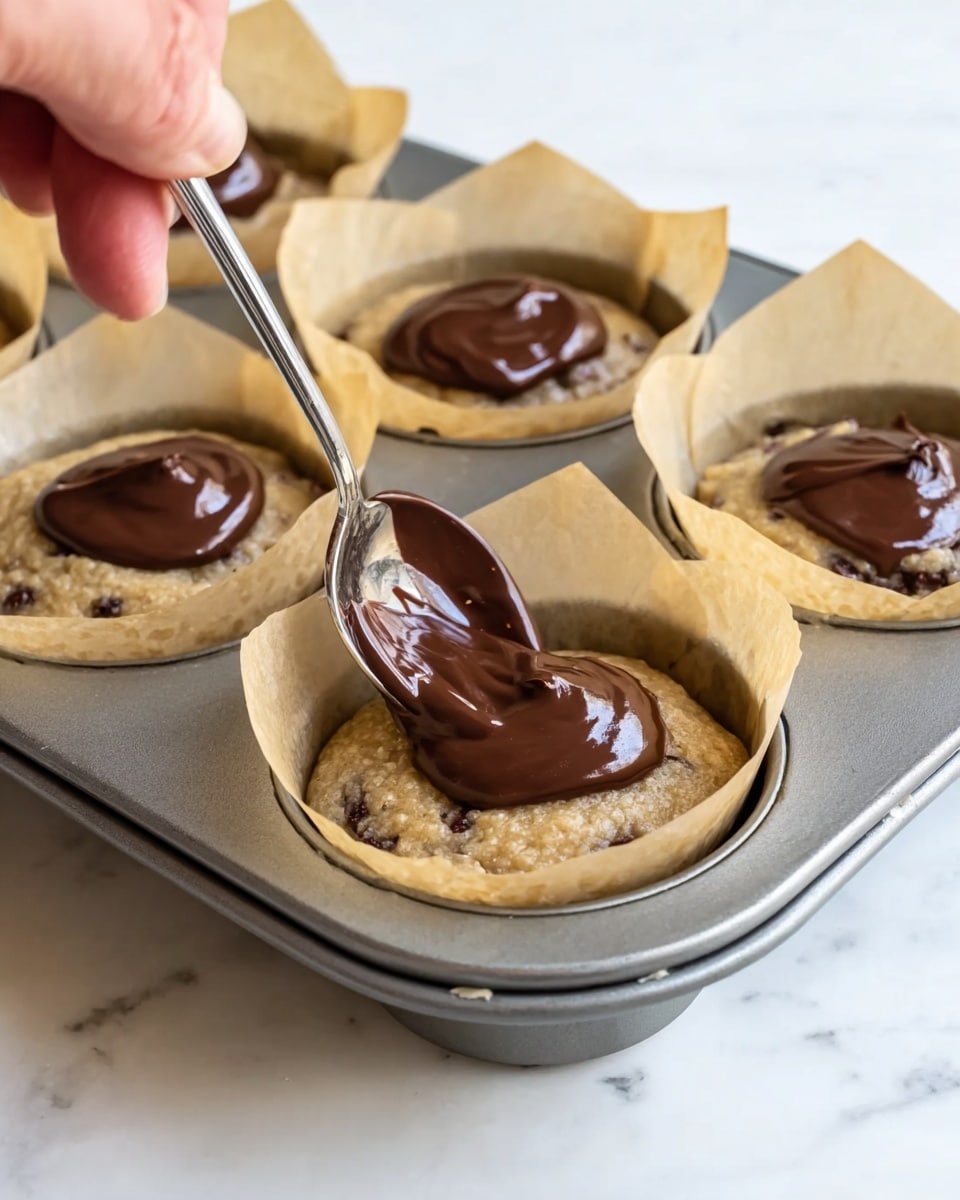 This image shows a metal muffin tray holding four cups lined with beige parchment paper. Each cup is filled with light brown batter mixed with small dark spots, and a dollop of thick, dark brown chocolate sits on top of each batter layer. A woman's hand is holding a silver spoon above the closest cup, spreading the chocolate on the batter. The background is a white marbled surface. photo taken with an iphone --ar 4:5 --v 7