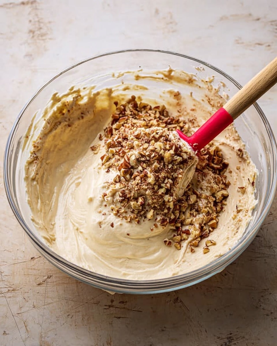 A clear glass bowl sits on a white marbled surface filled with creamy, light beige batter that has a smooth and thick texture. On top of the batter, there is a large sprinkle of finely chopped brown nuts, mainly spread near the center but blending into the batter on the sides. A wooden spatula with a red silicone tip rests inside the bowl, partially covered with batter. The bowl shows some batter wiped around its inside edges. Photo taken with an iphone --ar 4:5 --v 7