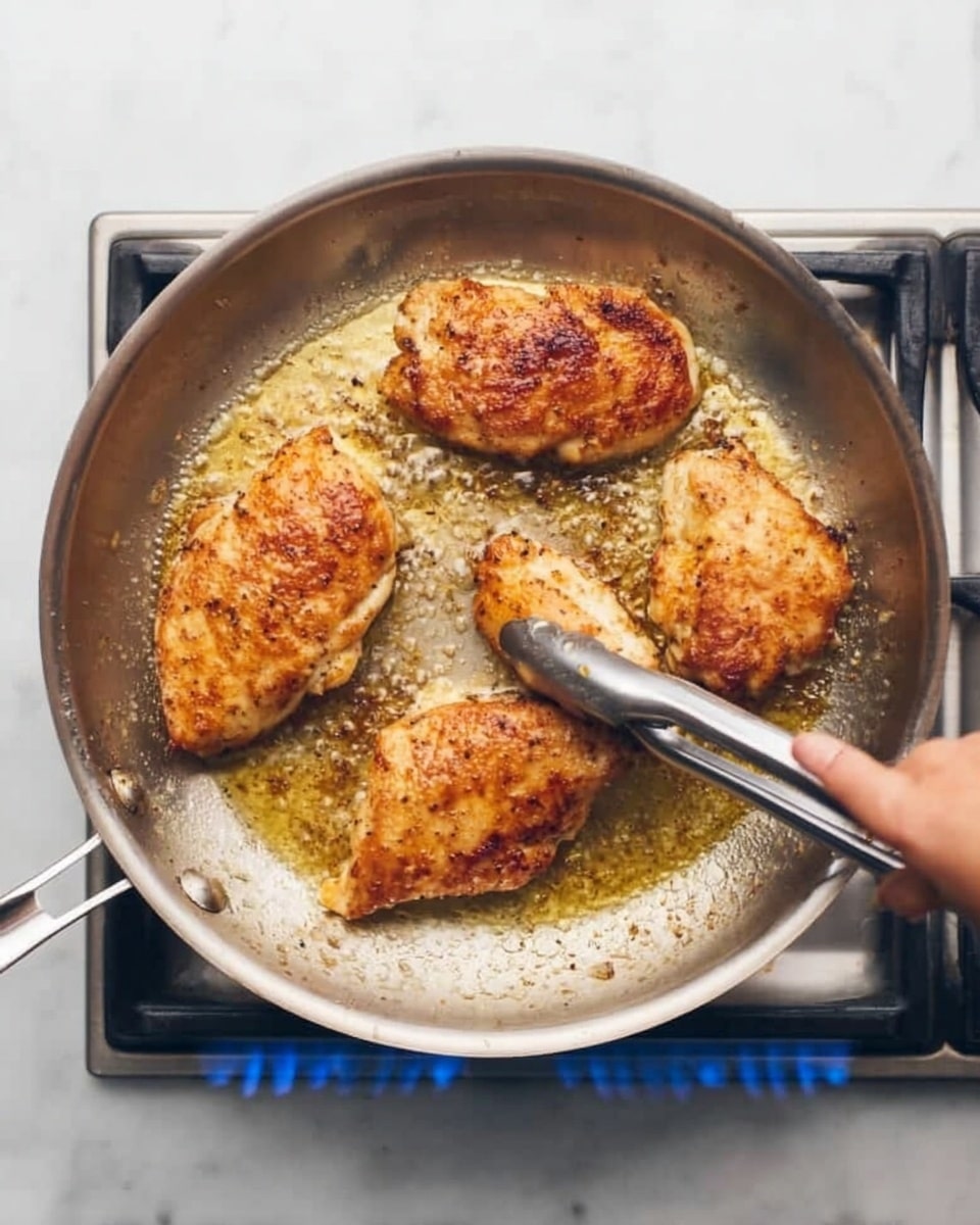 Four golden brown pieces of chicken are cooking in a shiny silver pan on top of a gas stove with blue flames visible below. A woman's hand is holding silver tongs picking up one piece, which shows a textured, crispy surface with light browning. The pan has melted butter or oil bubbling around the chicken, creating a glossy finish. The stove is set on a clean white marbled surface. photo taken with an iphone --ar 4:5 --v 7