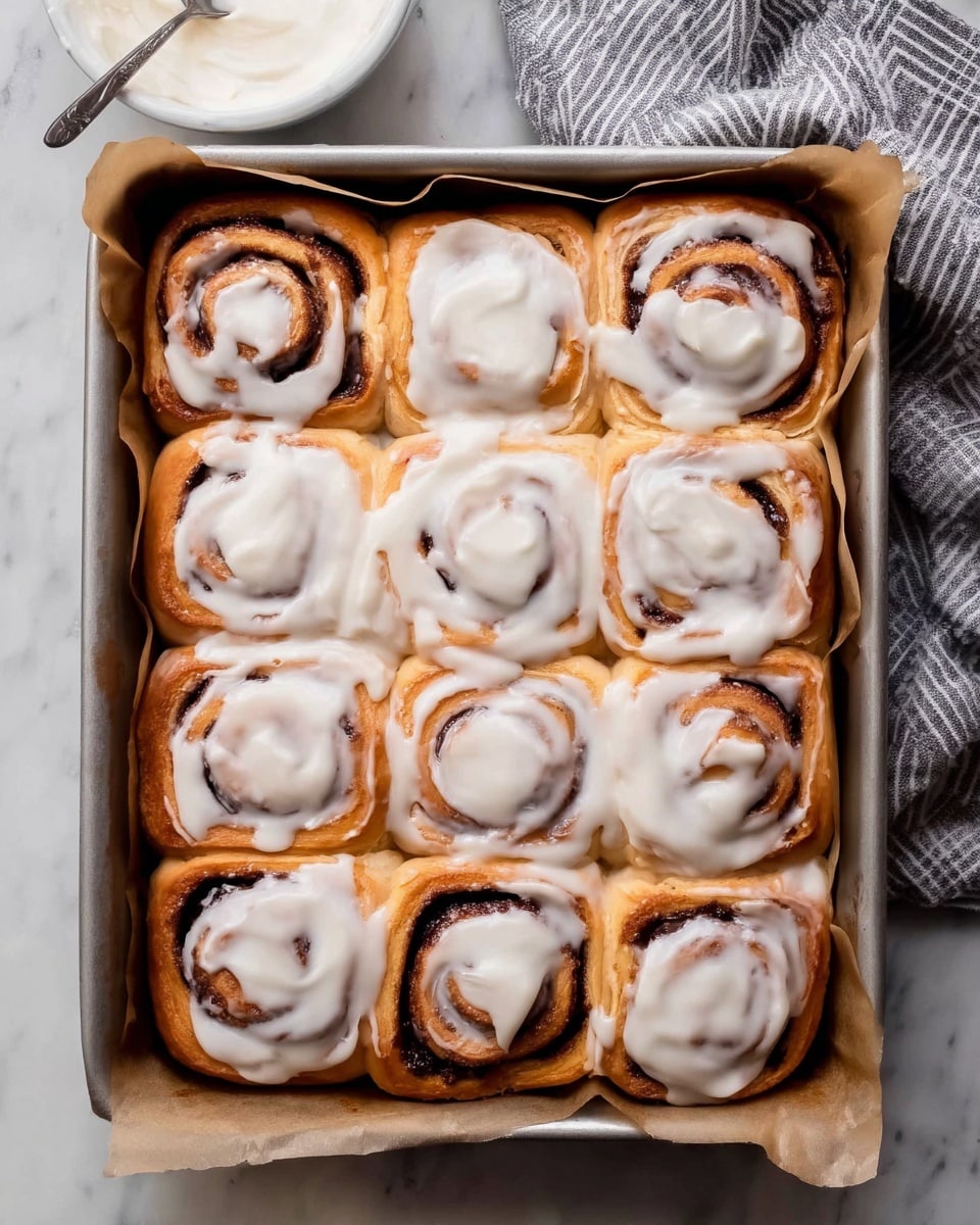 A metal baking pan lined with brown paper holds twelve cinnamon rolls arranged in a 3 by 4 grid. Each cinnamon roll is golden-brown with dark brown swirls of cinnamon filling visible through the spiraled dough. The rolls are generously covered with a smooth, thick white icing that drapes over the tops and seeps into the spaces between the rolls, giving a glossy, creamy texture. In the bottom part of the image, a white bowl filled with the same white icing sits on a white marbled surface. A gray and white striped cloth is partially visible at the top right corner. photo taken with an iphone --ar 4:5 --v 7