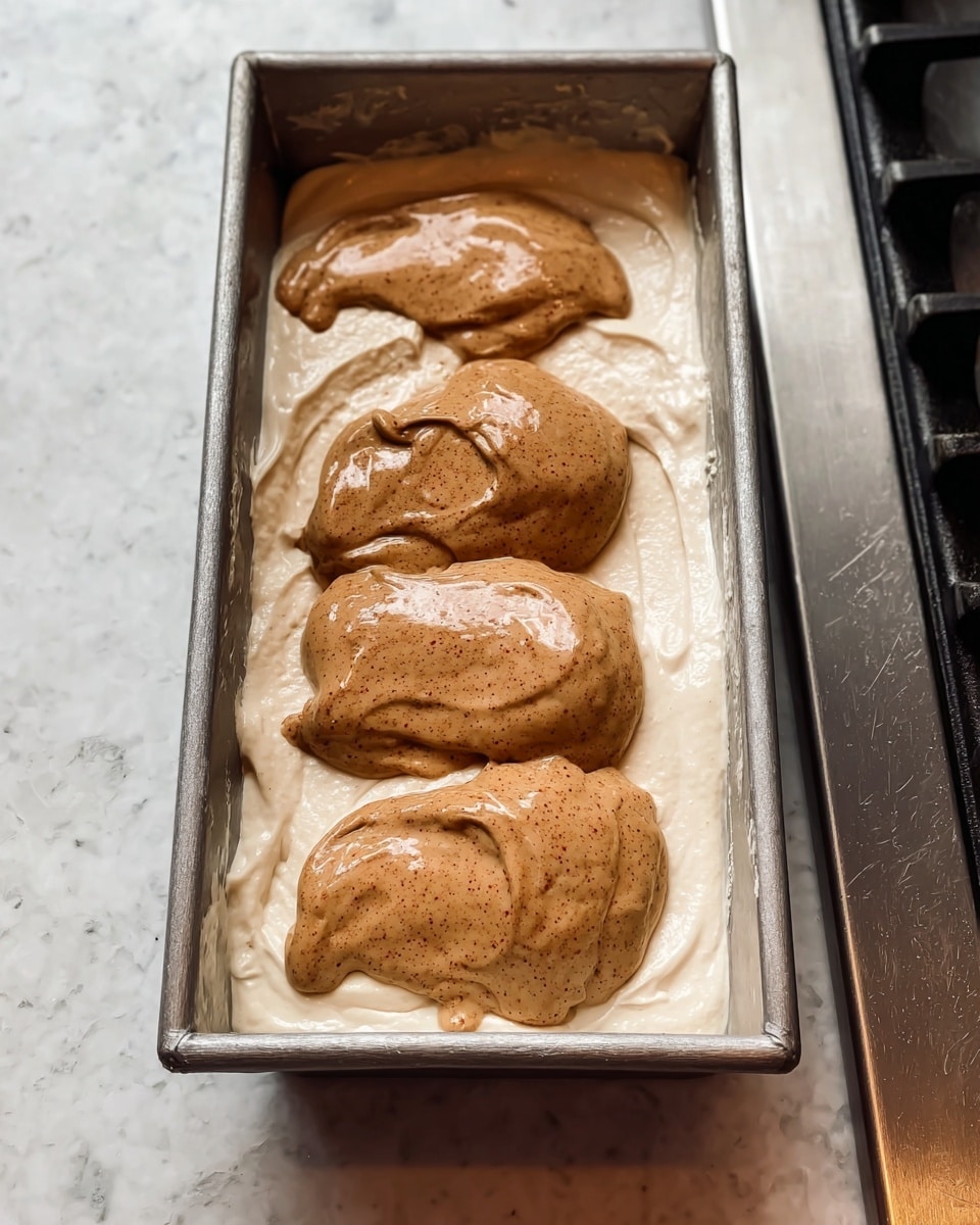 A rectangular metal baking pan holds two layers of batter. The bottom layer is smooth, thick, and white. On top, there are four uneven dollops of a light brown batter with a shiny, slightly speckled texture, placed in a row along the length of the pan. The pan is resting on a silver stovetop with a white marbled surface around it. photo taken with an iphone --ar 4:5 --v 7