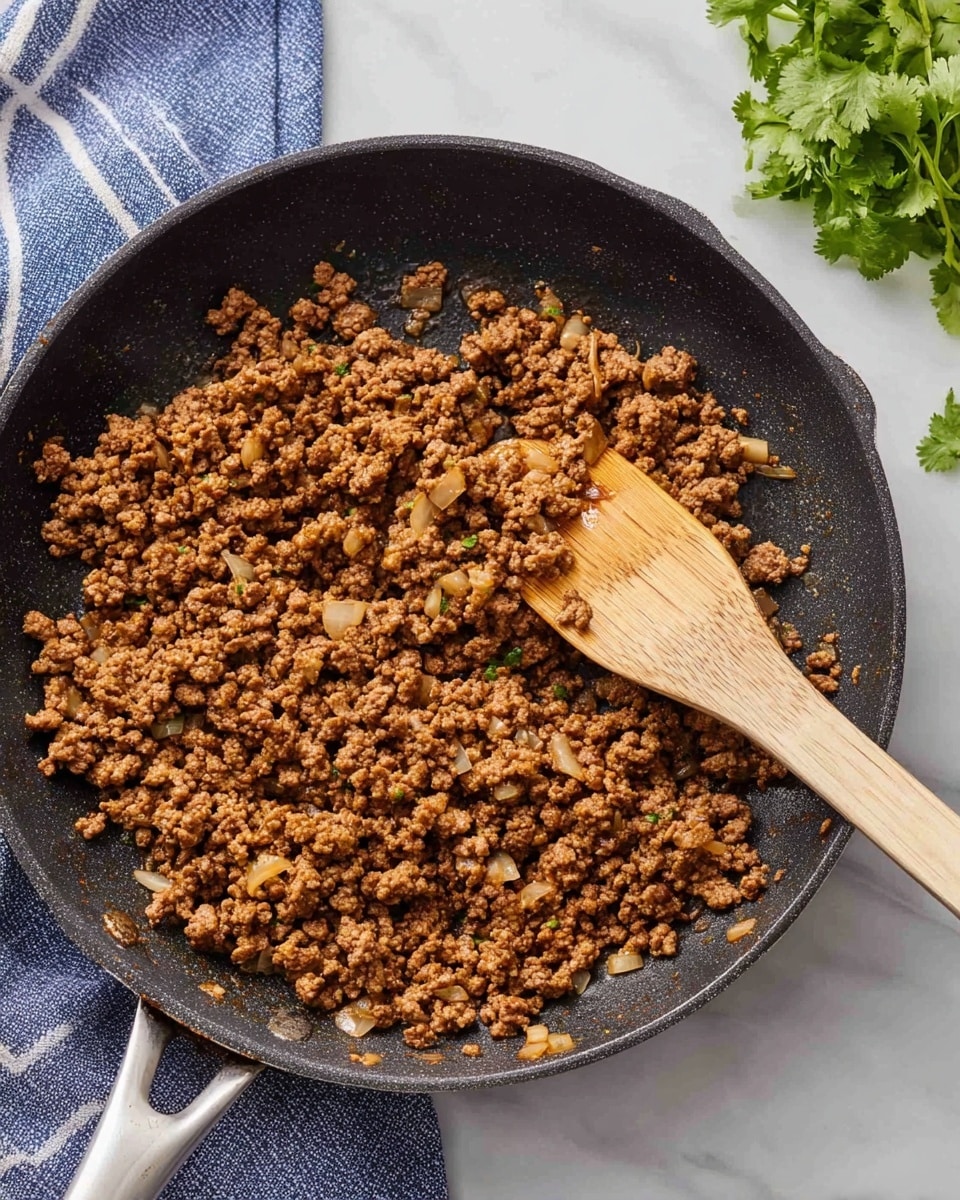 A black frying pan filled with browned ground meat mixed with small pieces of cooked onions, showing a crumbly texture. A wooden spatula is resting inside the pan, partially lifting some of the meat mixture. The pan handle is visible on the right side. The pan sits on a white marbled surface with a blue and white striped cloth nearby and some green cilantro leaves at the top. Photo taken with an iphone --ar 4:5 --v 7
