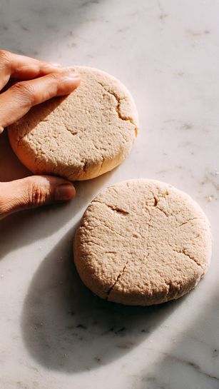 The image shows two round, flat dough pieces with a light beige color and a slightly rough texture. They have small cracks on the surface, indicating softness and pliability. The dough pieces are placed on a white marbled surface, spaced apart. The lighting is soft, highlighting the dough's natural color and texture. In the bottom left corner, a woman's hand is about to touch one of the dough pieces. Photo taken with an iphone --ar 4:5 --v 7