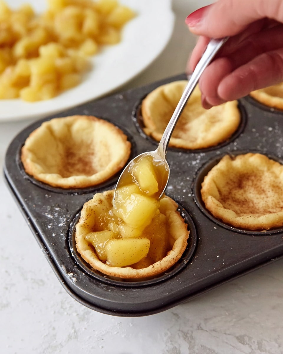 A dark metal muffin tray with six small, round crust shells placed inside each slot is shown on a white marbled surface. Each crust is light golden with a slightly uneven texture and has visible cinnamon specks spread throughout. A woman's hand is holding a silver spoon filled with shiny, golden-yellow apple filling with soft fruit chunks, poised over one of the crust shells ready to add the filling. In the blurred background, a white plate holds more apple filling. Photo taken with an iphone --ar 4:5 --v 7