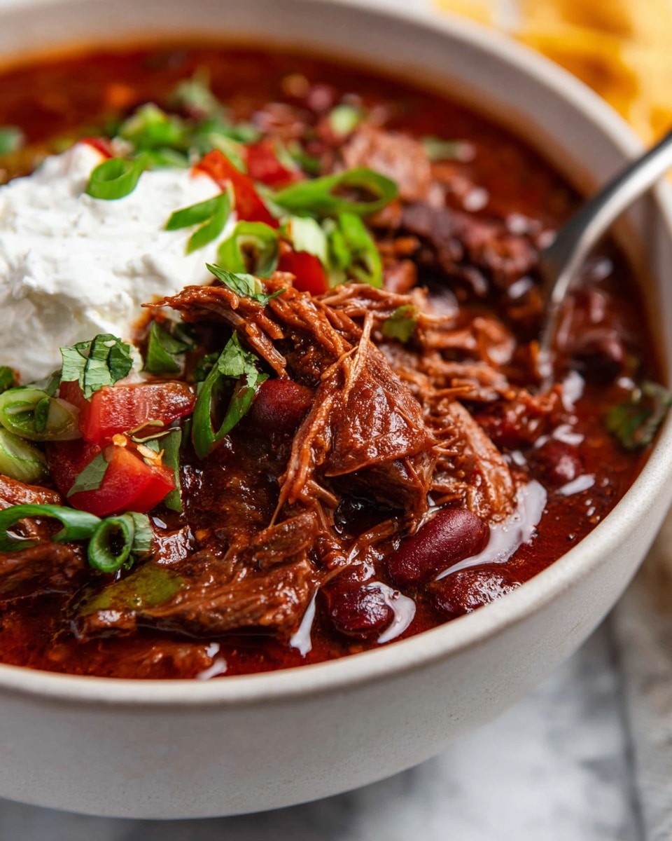The dish is a bowl filled with rich, dark red chili with chunks of tender shredded meat layered throughout. On top, there are fresh green slices of jalapeño peppers, chopped green onions, and thin shreds of orange cheddar cheese. A dollop of white sour cream sits near the center, surrounded by small scattered cilantro leaves. The bowl is white and placed on a white marbled surface with some cilantro sprigs and a broken piece of tortilla chip nearby. The texture of the chili looks thick and hearty with visible kidney beans mixed in. photo taken with an iphone --ar 4:5 --v 7