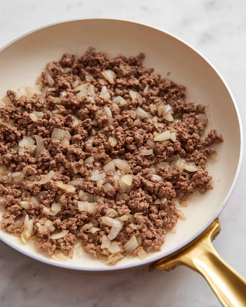 The image shows a white frying pan filled with cooked ground beef mixed with soft, translucent chopped onions spread evenly across the pan's base. The beef appears crumbly with a brown color, mixed closely with pale yellowish white onion pieces, creating a textured, hearty look. The frying pan has a shiny gold handle and is placed on a white marbled surface, with soft natural light highlighting the details. photo taken with an iphone --ar 4:5 --v 7