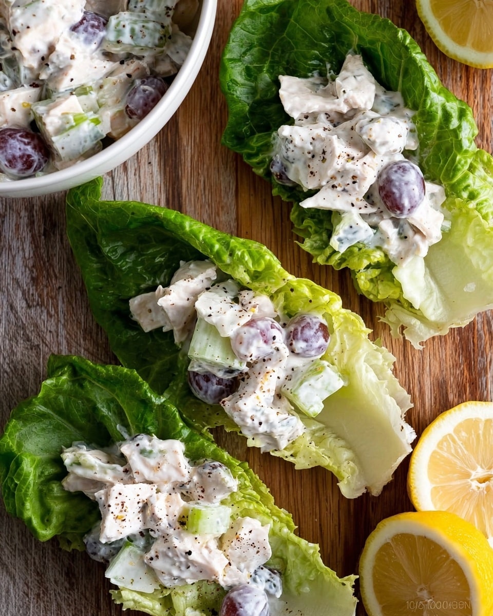 The image shows three green lettuce leaves laid flat on a wooden surface, each holding a creamy white salad mix. The salad contains visible pieces of white chicken, celery, and purple grapes, all coated in a thick, white creamy dressing with some black pepper sprinkled on top. In the top left corner, part of a white bowl filled with the same salad is visible, and on the right side are two halves of a lemon with one facing up and the other down, showing the juicy pulp inside. photo taken with an iphone --ar 4:5 --v 7
