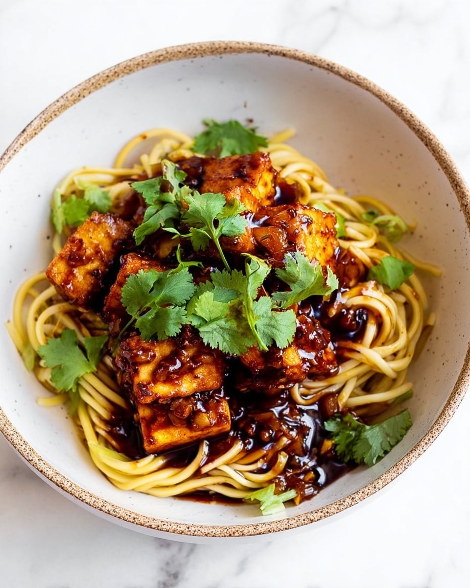 A white bowl with a speckled rim holds a dish of light yellow noodles at the base, topped with several pieces of golden brown, glazed fried tofu stacked in the center. Bright green cilantro leaves are scattered over the tofu, adding fresh color contrast. The tofu has a glossy, thick dark sauce coating it, which drips slightly onto the noodles. The bowl sits on a white marbled surface. photo taken with an iphone --ar 4:5 --v 7