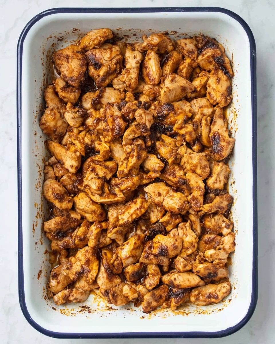 A white rectangular baking dish with a dark blue rim filled with many pieces of cooked chicken, all with a browned and slightly charred texture. The chicken pieces are small and uneven in shape, layered closely covering the entire dish. The chicken looks seasoned with spices giving a warm brown and orange color. The dish sits on a white marbled surface. Photo taken with an iphone --ar 4:5 --v 7