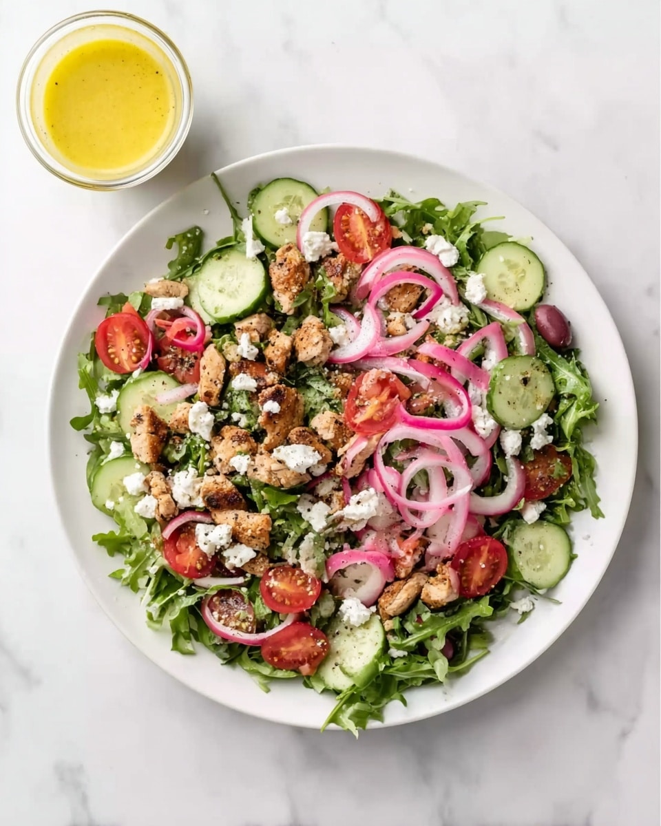 A white plate with a fresh salad sits on a white marbled surface. The salad has a bottom layer of green leafy arugula, topped with scattered pieces of cooked brown chicken, thinly sliced cucumber rounds, halved red cherry tomatoes, and thin rings of pink pickled red onions. Small white chunks of feta cheese are spread evenly on top. To the upper left of the plate, there is a small clear glass container filled with yellow salad dressing. The photo taken with an iphone --ar 4:5 --v 7