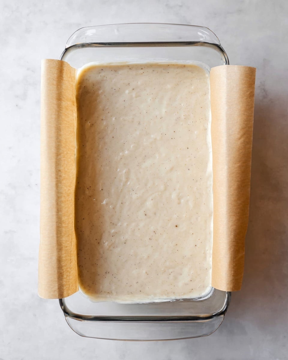 The image shows a clear glass rectangular dish lined with light brown parchment paper folded over the shorter sides. Inside the dish is a smooth, creamy batter layer that is light beige with small dark specks evenly spread throughout. The dish sits on a white marbled surface that contrasts softly with the batter color. The surface of the batter is slightly uneven with small bubbles and gentle texture. photo taken with an iphone --ar 4:5 --v 7