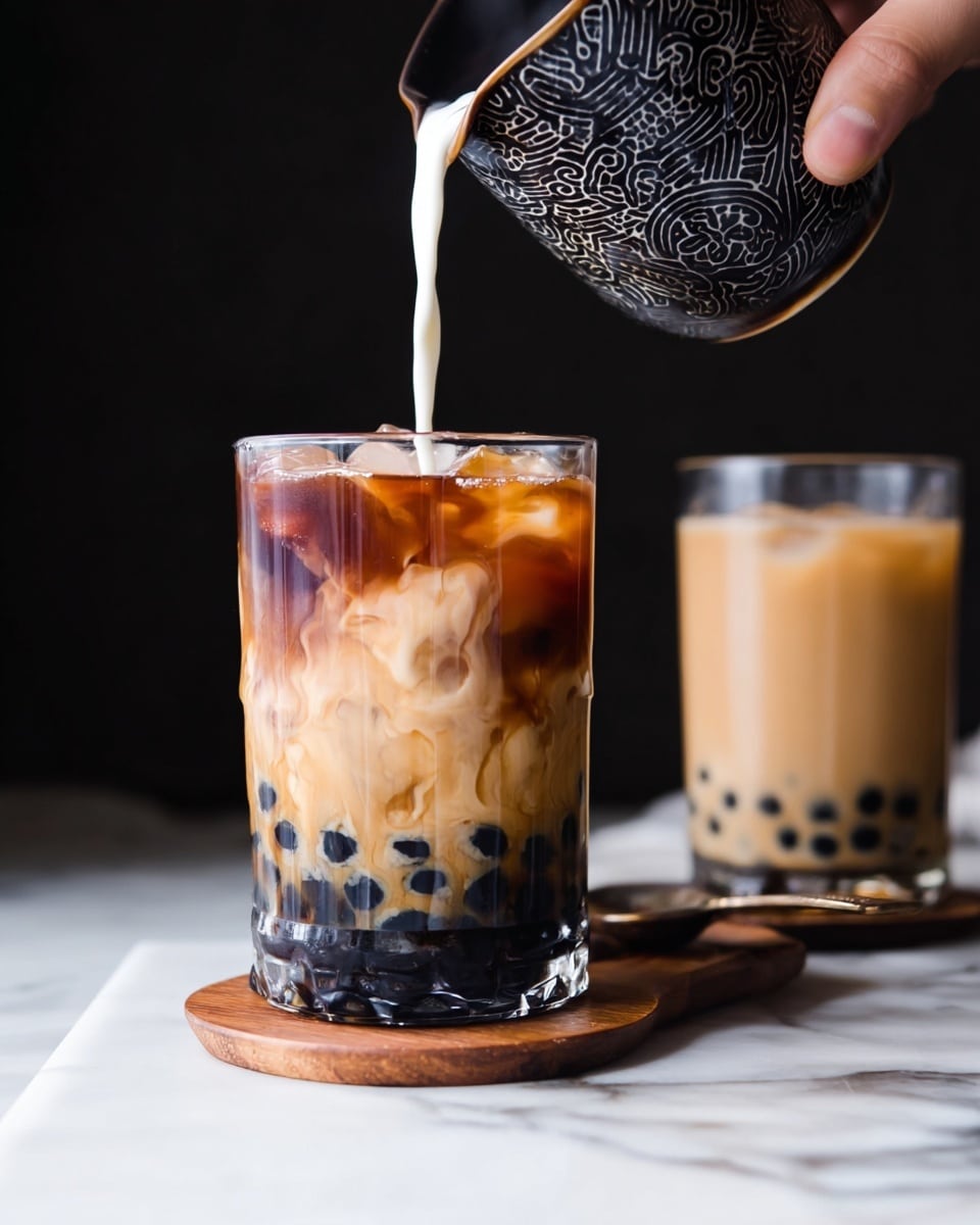 A clear glass filled with black tapioca pearls at the bottom, a dark brown layer of iced tea above the pearls, and light cream being poured from a patterned black cup, swirling softly into the tea creating creamy beige marbled patterns; next to it is another clear glass of creamy, light brown bubble tea with black tapioca pearls at the bottom, placed on a wooden coaster, all set on a white marbled surface with a black background, a woman's hand holding the cup pouring cream photo taken with an iphone --ar 4:5 --v 7