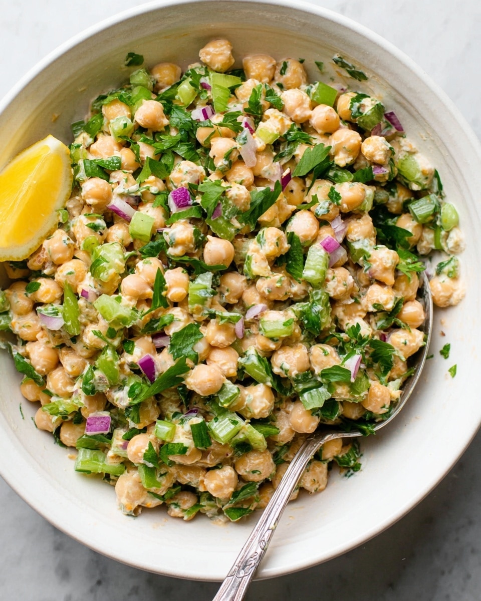 The image shows a white bowl filled with a mixed salad. The main layer inside the bowl is made of small, round chickpeas with a creamy dressing giving them a soft, pale yellow color. Scattered evenly throughout are pieces of bright green celery and small cubes of purple-red onions. Fresh parsley leaves are sprinkled on top, adding fresh dark green accents. A wedge of yellow lemon sits at the edge of the bowl. A silver spoon rests partially buried in the mixture, all set against a white marbled background. Photo taken with an iphone --ar 4:5 --v 7