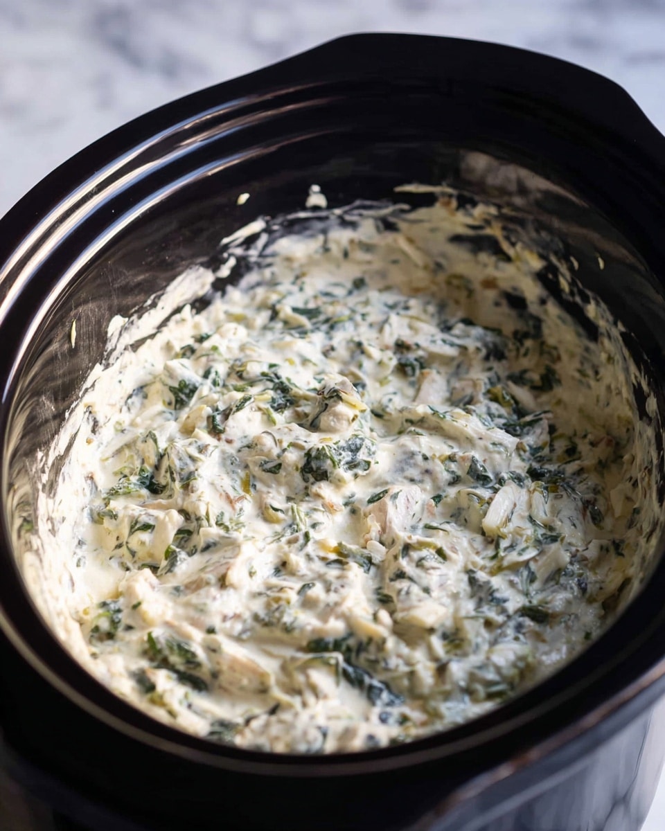 A black slow cooker filled with a thick, creamy dip that has a white base mixed with green leafy pieces and light beige chunks spread evenly throughout. The dip has a slightly lumpy texture with visible bits of herbs and vegetables. The slow cooker sits on a white marbled surface, and the interior rim of the cooker is visible with some dip residue. The lighting is natural and soft, highlighting the creamy texture of the dish. photo taken with an iphone --ar 4:5 --v 7