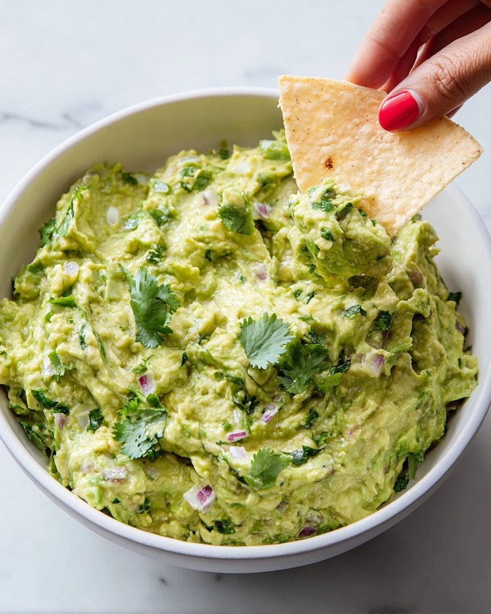 A white bowl filled with creamy green guacamole mixed with small bits of red onion and green herbs, with a slightly chunky texture and some fresh cilantro leaves scattered on top. A woman's hand with red nail polish holds a triangular, light beige tortilla chip dipping into the guacamole on the right side. The background is a white marbled surface. photo taken with an iphone --ar 4:5 --v 7