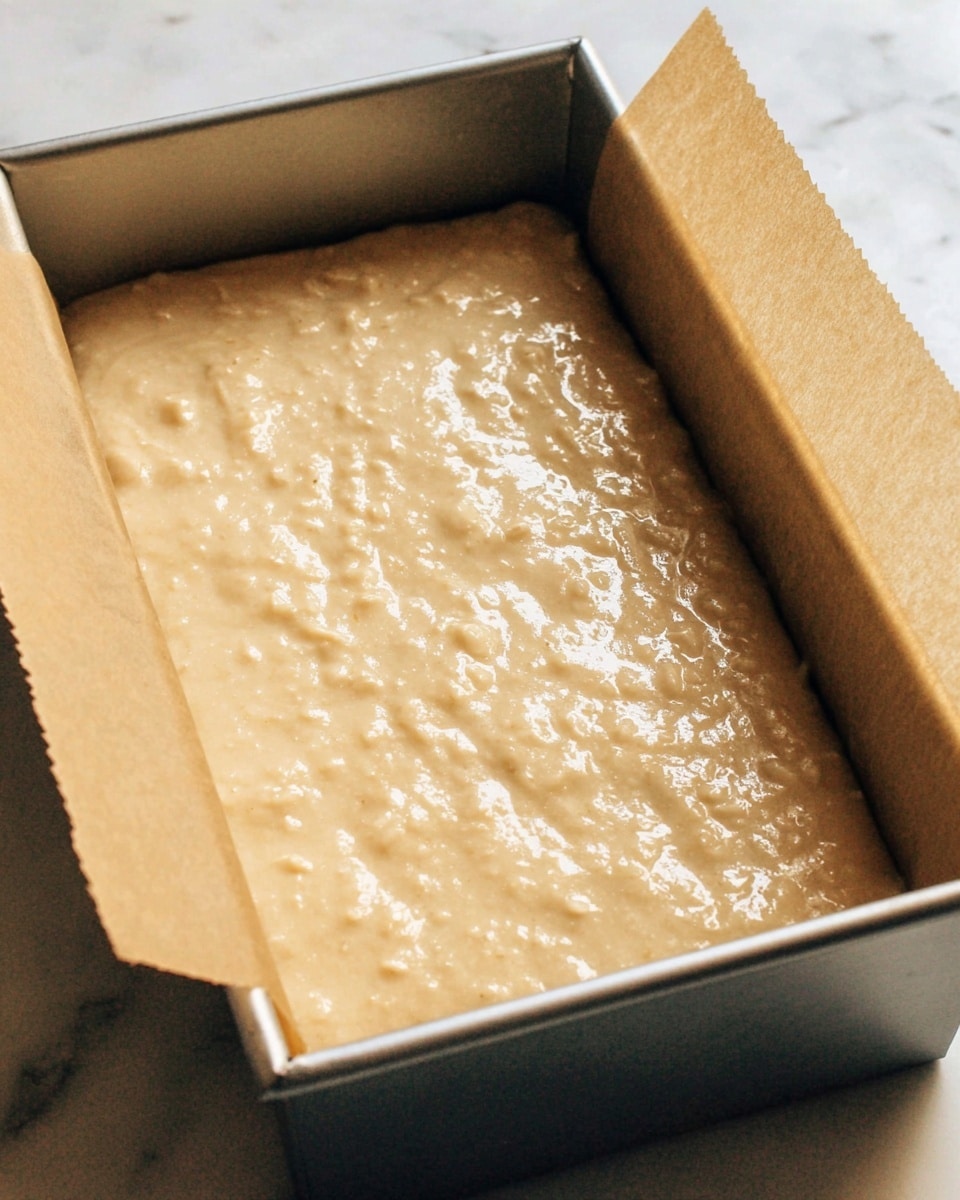 A close-up view of a rectangular metal baking pan lined with light brown parchment paper, filled with a smooth, thick batter that is light beige with small lumps and a slightly glossy texture, evenly spread to fill the pan. The background is a white marbled texture. photo taken with an iphone --ar 4:5 --v 7