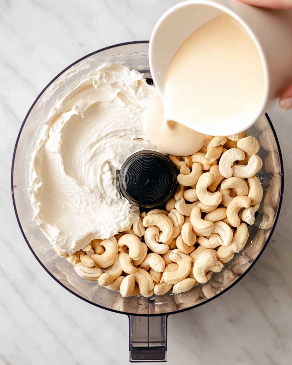 A bird's eye view of a clear food processor bowl containing three layers: the bottom layer is light beige cashew nuts, whole and curved; the middle layer is thick, white creamy substance spread unevenly around the nuts; the top layer shows a woman's hand pouring a light beige liquid from a white cup into the bowl. The bowl is placed on a white marbled surface photo taken with an iphone --ar 4:5 --v 7