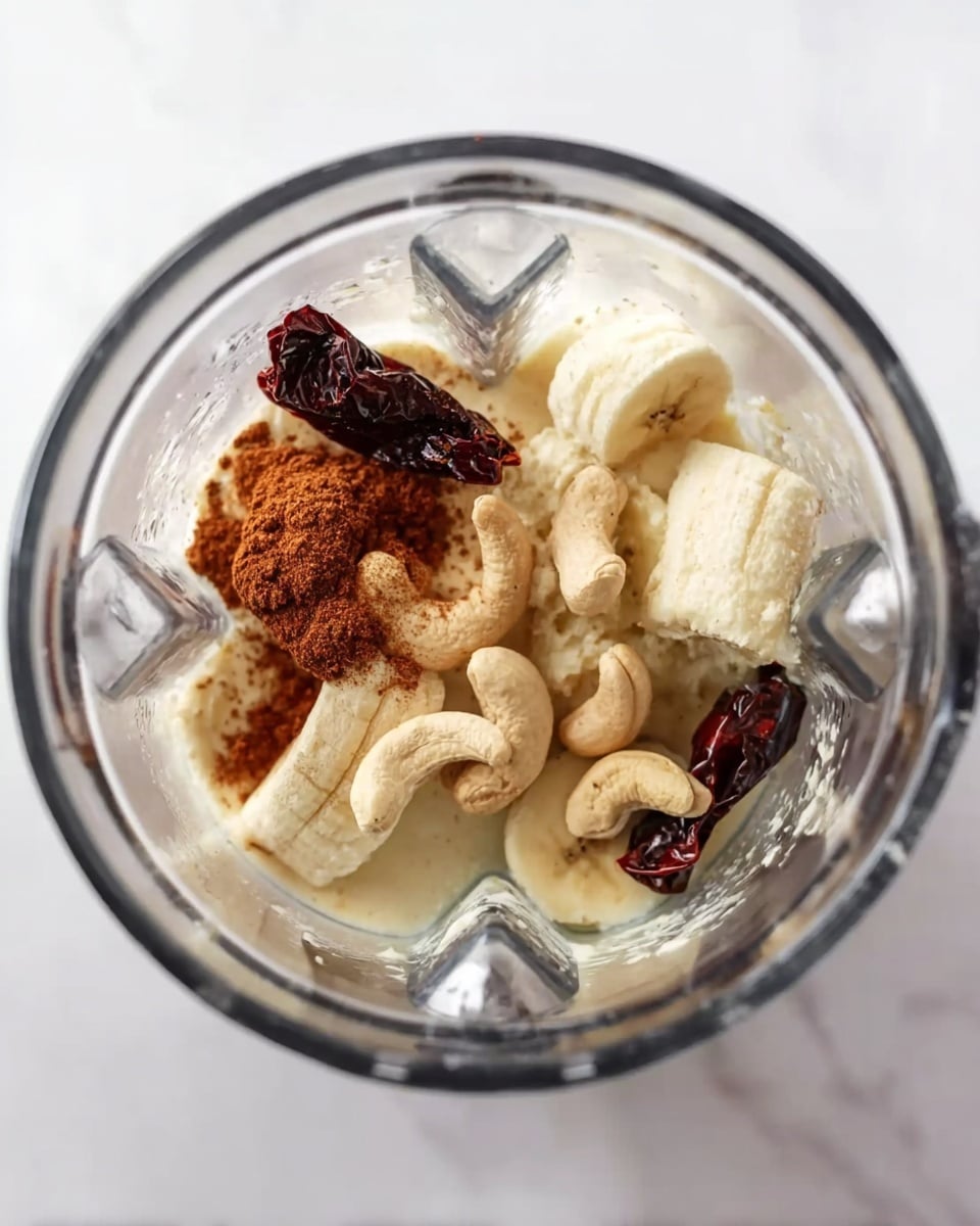 The image shows a close-up top view of a clear glass blender filled with ingredients. At the bottom, there are thick white banana slices with some creamy light beige yogurt around them. Scattered on top are several whole light brown cashew nuts and a light dusting of brown cinnamon powder. Dark reddish-brown dried chili peppers are placed a few times around the blender, contrasting with the lighter ingredients beneath. The clear glass blender is placed on a white marbled surface. photo taken with an iphone --ar 4:5 --v 7