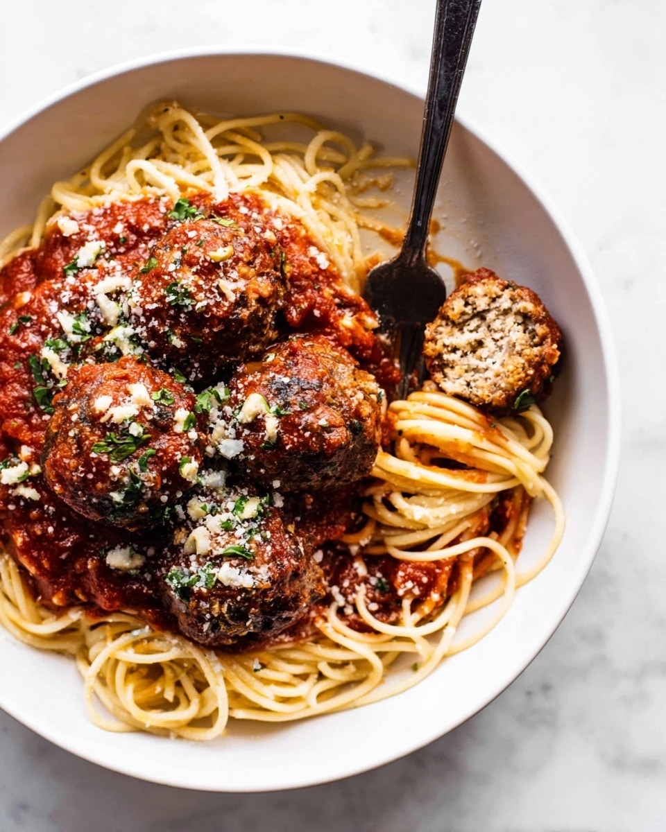A white bowl holds a bed of light yellow spaghetti noodles, loosely piled in the bottom half and sides, with a thick dark red tomato sauce covering parts of the noodles. Four brown meatballs sit on top, each showing a rough, textured surface with some herbs sprinkled over them. One meatball is cut in half, revealing a dense, mixed interior with bits of mushroom visible. Small bits of white cheese and green herbs are sprinkled on top of the meatballs and sauce. A dark metal fork is placed on the right side of the bowl, with the tines resting in the partially cut meatball. The bowl is set on a white marbled surface. Photo taken with an iphone --ar 4:5 --v 7