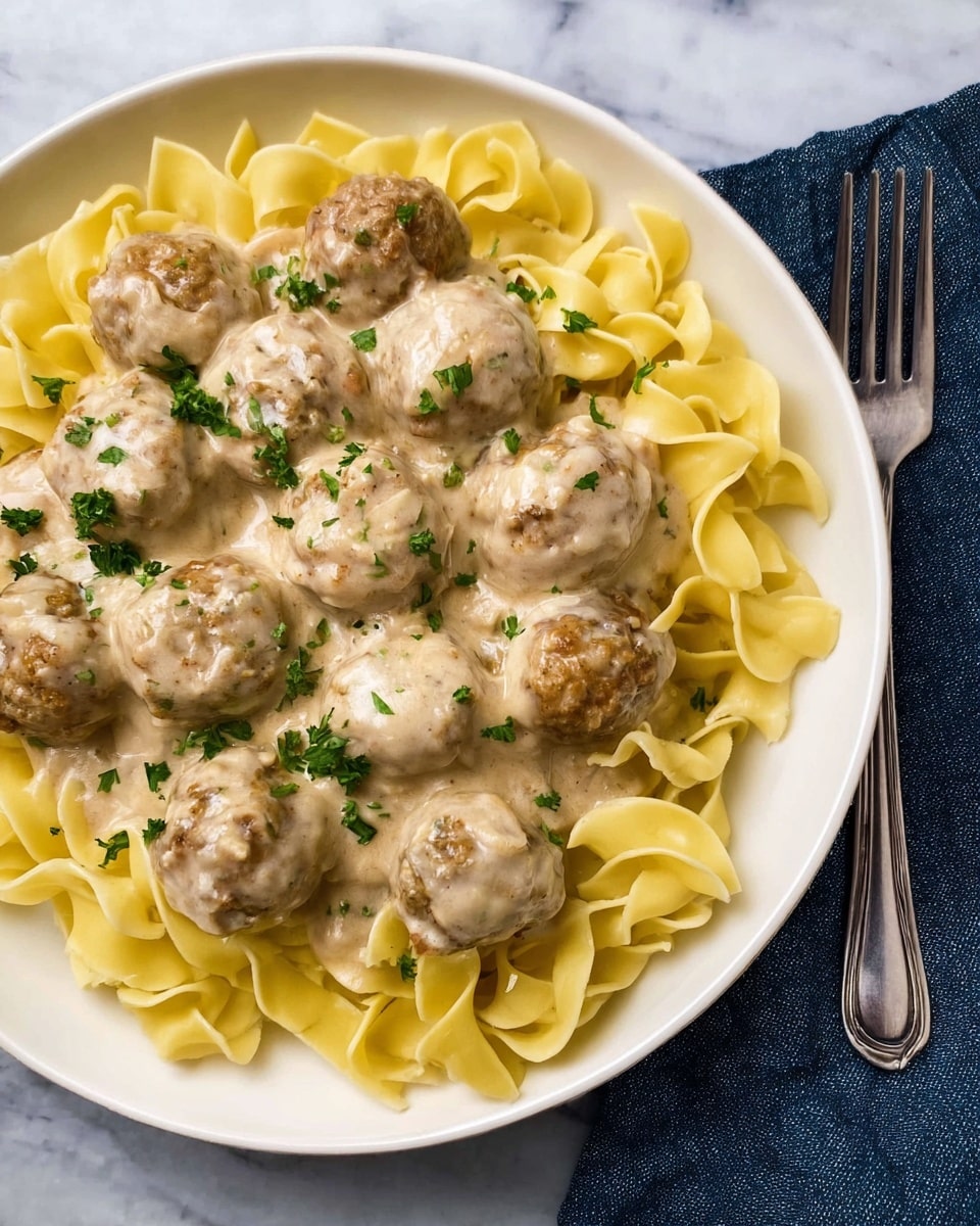 A white plate holds a bed of light yellow wide egg noodles with soft wavy texture arranged in a loose pile around the edge. On top, there are about fourteen round brown meatballs covered in a thick light beige creamy sauce that looks smooth with some specks of seasoning. Small green parsley pieces are scattered on top of the sauce and meatballs, adding color contrast. The plate rests on a white marbled surface, with a silver fork and dark blue cloth to the side. photo taken with an iphone --ar 4:5 --v 7