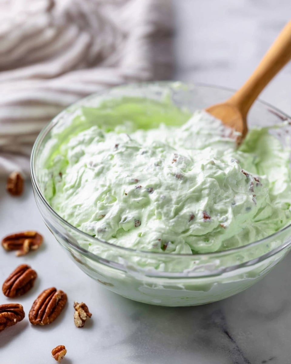 A clear glass bowl filled with a fluffy, light green creamy mixture with visible small chunks mixed throughout. The texture looks soft and thick with a slightly lumpy surface. A wooden spoon with a cream-covered tip rests in the bowl, leaning against the side. The bowl sits on a white marbled surface with several whole and broken pecan nuts scattered nearby. A striped cloth is partially visible in the soft-focus background. Photo taken with an iphone --ar 4:5 --v 7