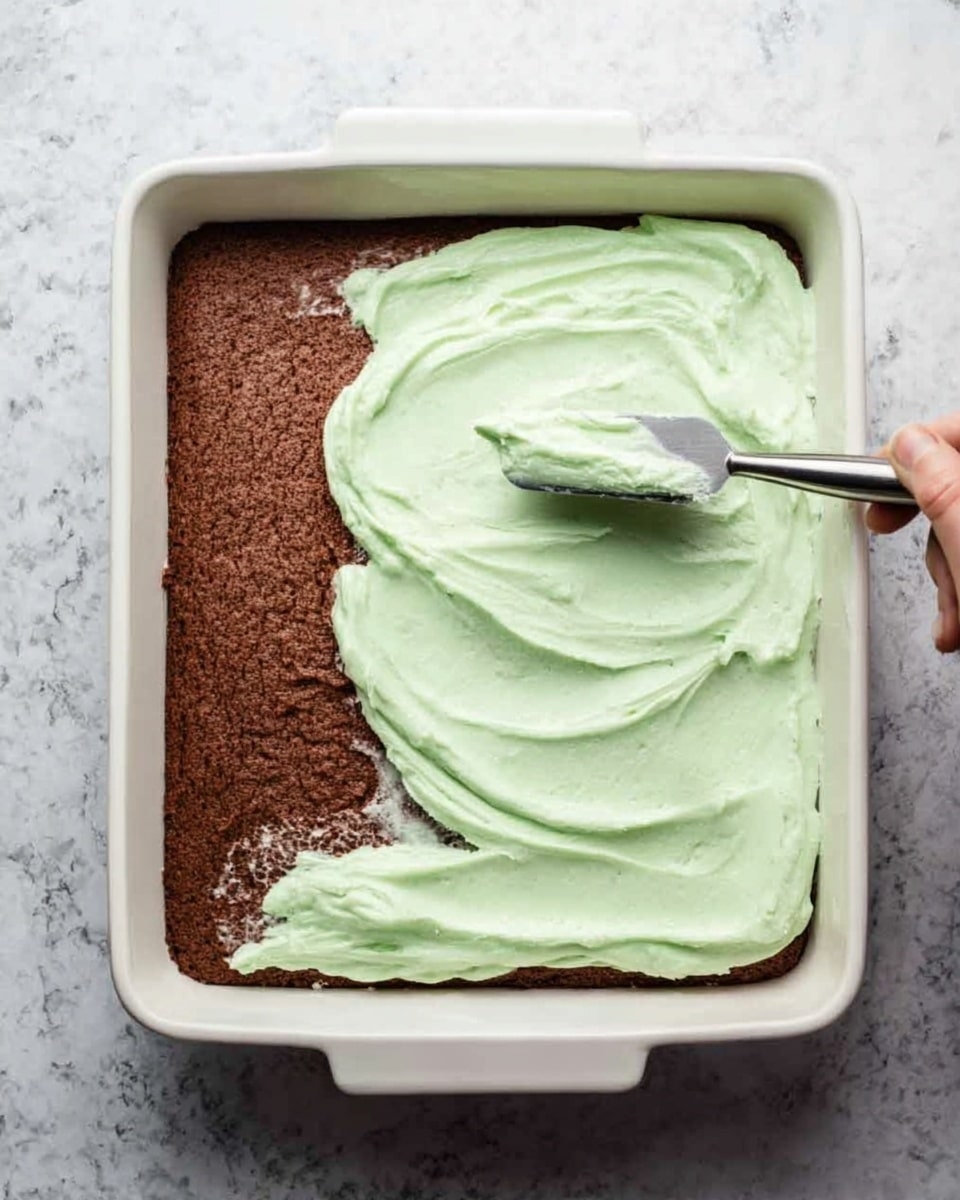 A white rectangular baking dish filled partly with a brown cake base on the right side and a thick layer of smooth, light green frosting being spread over the left side of the cake with a metal spatula held by a woman's hand. The frosting has a creamy texture and covers about half of the cake, with the spatula in motion creating soft swirls. The dish is placed on a white marbled textured surface. photo taken with an iphone --ar 4:5 --v 7