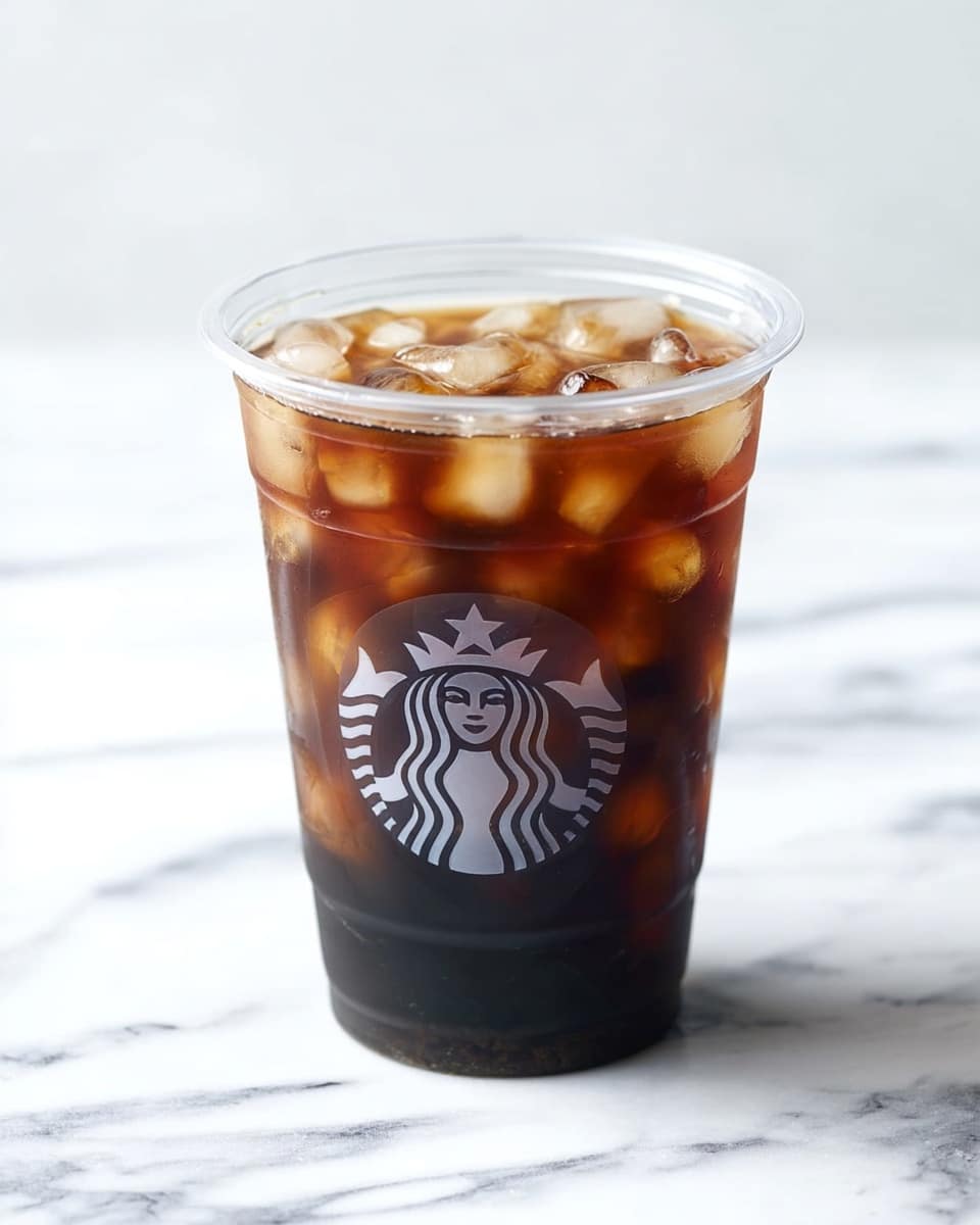 A clear plastic cup with the Starbucks logo is filled with dark brown cold brew coffee and light tan ice cubes floating on top and throughout the drink, creating a mix of dark and lighter brown shades. The cup is placed on a white marbled surface with a blurred white background. Photo taken with an iphone --ar 4:5 --v 7