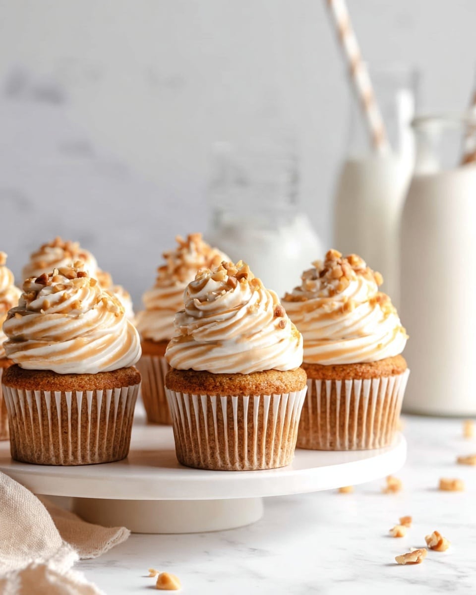 This image shows five cupcakes placed on a round white stand on a white marbled surface. Each cupcake has a golden brown base with ridges from the cupcake liner. On top of each cupcake is a swirl of smooth, light cream-colored frosting, layered in a spiral shape that forms a peak. Small pieces of chopped nuts are sprinkled over the frosting, adding texture and a touch of light brown color. In the background, there are two glass bottles with straws and a white container, all slightly out of focus. Scattered nut pieces are also visible around the stand. Photo taken with an iphone --ar 4:5 --v 7