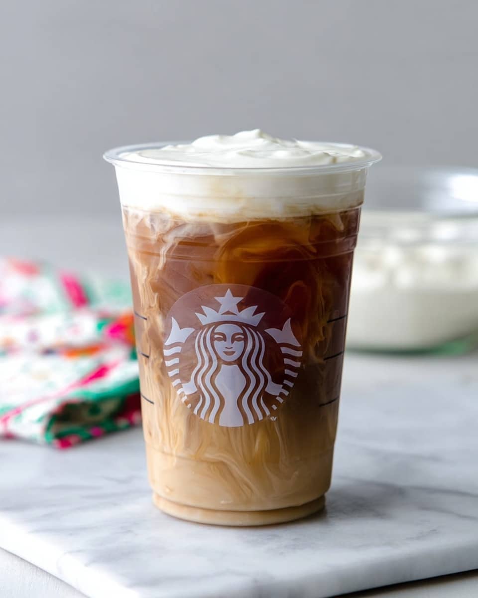 A clear plastic cup filled with a three-layer iced coffee drink sits on a white marbled surface. The bottom layer is a light brown coffee base with a slightly translucent texture. The middle layer is a darker brown coffee mix, showing some swirling lines where it blends with the bottom layer. The top layer is a thick, creamy white foam that rises above the rim of the cup with soft peaks. The cup has the Starbucks logo printed in white on its front. In the blurred background, a glass bowl with a white creamy substance is visible next to a colorful white cloth. Photo taken with an iphone --ar 4:5 --v 7