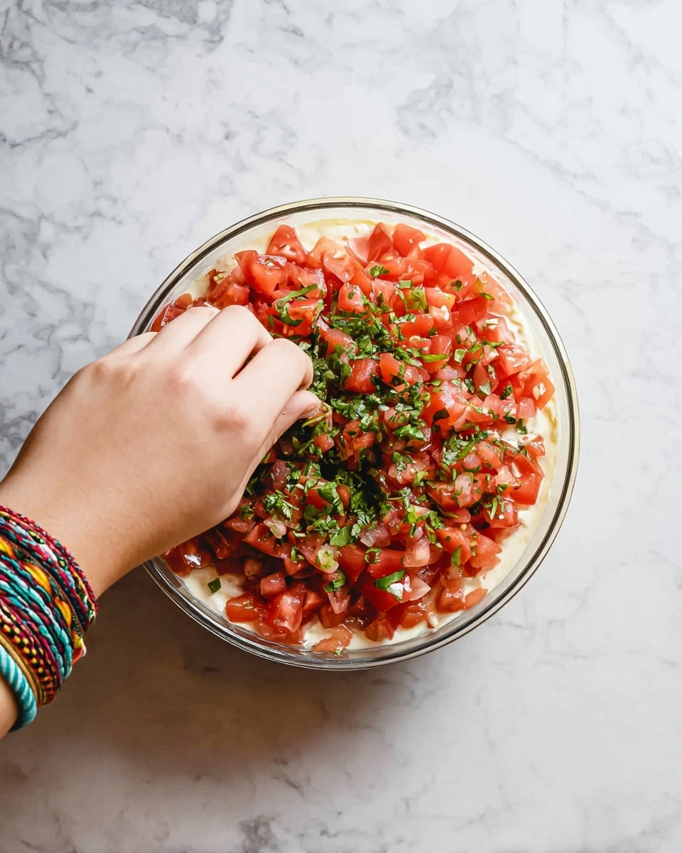 A clear round bowl with three visible layers is shown from above on a white marbled surface. The bottom layer is creamy and pale yellow, the middle layer is a bright white, and the top layer is covered with finely chopped red tomatoes mixed with small pieces of green herbs. A woman's hand wearing colorful woven bracelets reaches in from the left, placing or adjusting the herbs on the tomato layer. Photo taken with an iphone --ar 4:5 --v 7