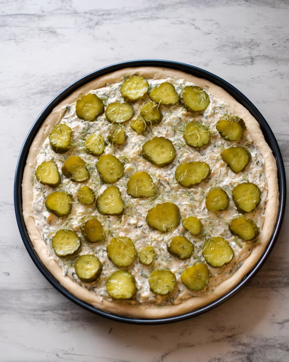 A round food item on a black pan placed on a white marbled surface. It has two visible layers: the bottom layer is a pale dough base, and the top layer is a spread of creamy white sauce with green herbs. On top, there are many round green pickle slices evenly spread out. The edges of the dough are slightly raised, holding the sauce and pickles inside. photo taken with an iphone --ar 4:5 --v 7