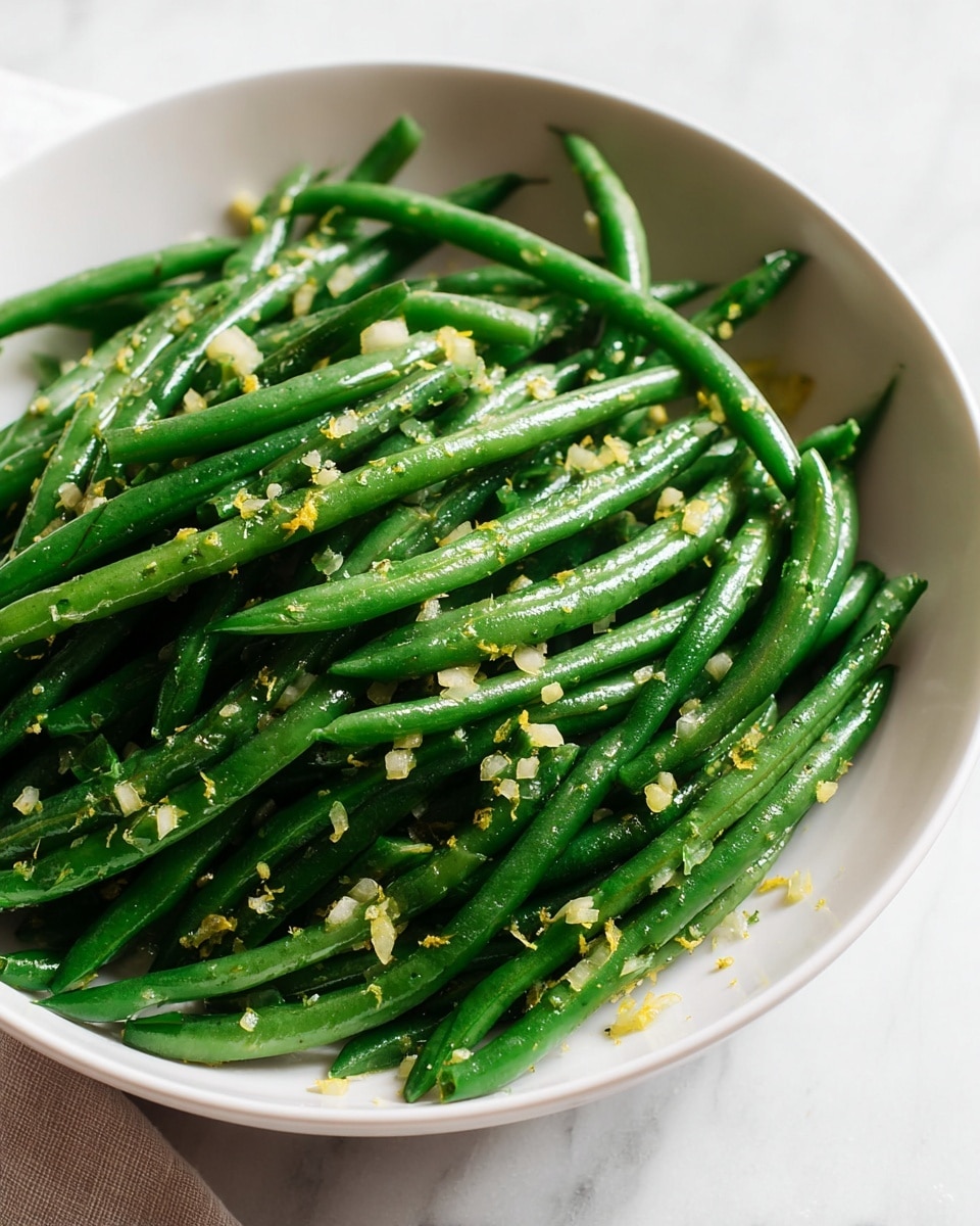 A white bowl holds a single layer of cooked green beans that are bright green and slightly shiny. The green beans are mixed with small pieces of finely chopped light-colored onion and tiny bits of yellow lemon zest. The dish looks fresh and light, resting on a white marbled surface with a soft, neutral background. Photo taken with an iphone --ar 4:5 --v 7