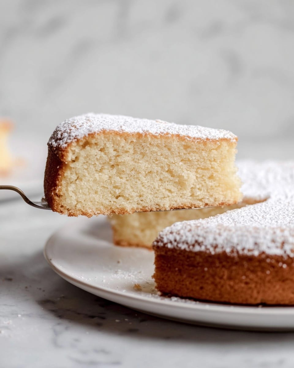A slice of soft, spongy cake is being lifted from a round cake on a white plate, showing its light, airy texture inside. The cake has a single thick layer, with a pale golden outer crust and a dusting of white powdered sugar on top. The background and surface are white marble texture, creating a clean and simple look. The focus is on the cake slice, highlighting its delicate crumb against the smooth powdered sugar top photo taken with an iphone --ar 4:5 --v 7