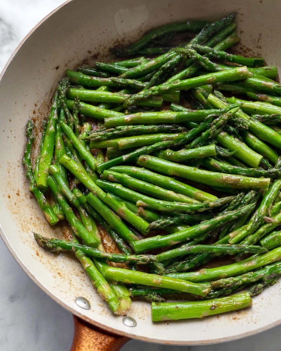 A close-up view of bright green asparagus spears cooked and placed inside a large off-white pan with a light brown handle. The asparagus is evenly spread in one layer, showing a shiny, slightly charred surface with small brown spots, suggesting it was cooked with oil or butter. The background features a white marbled texture. Photo taken with an iphone --ar 4:5 --v 7