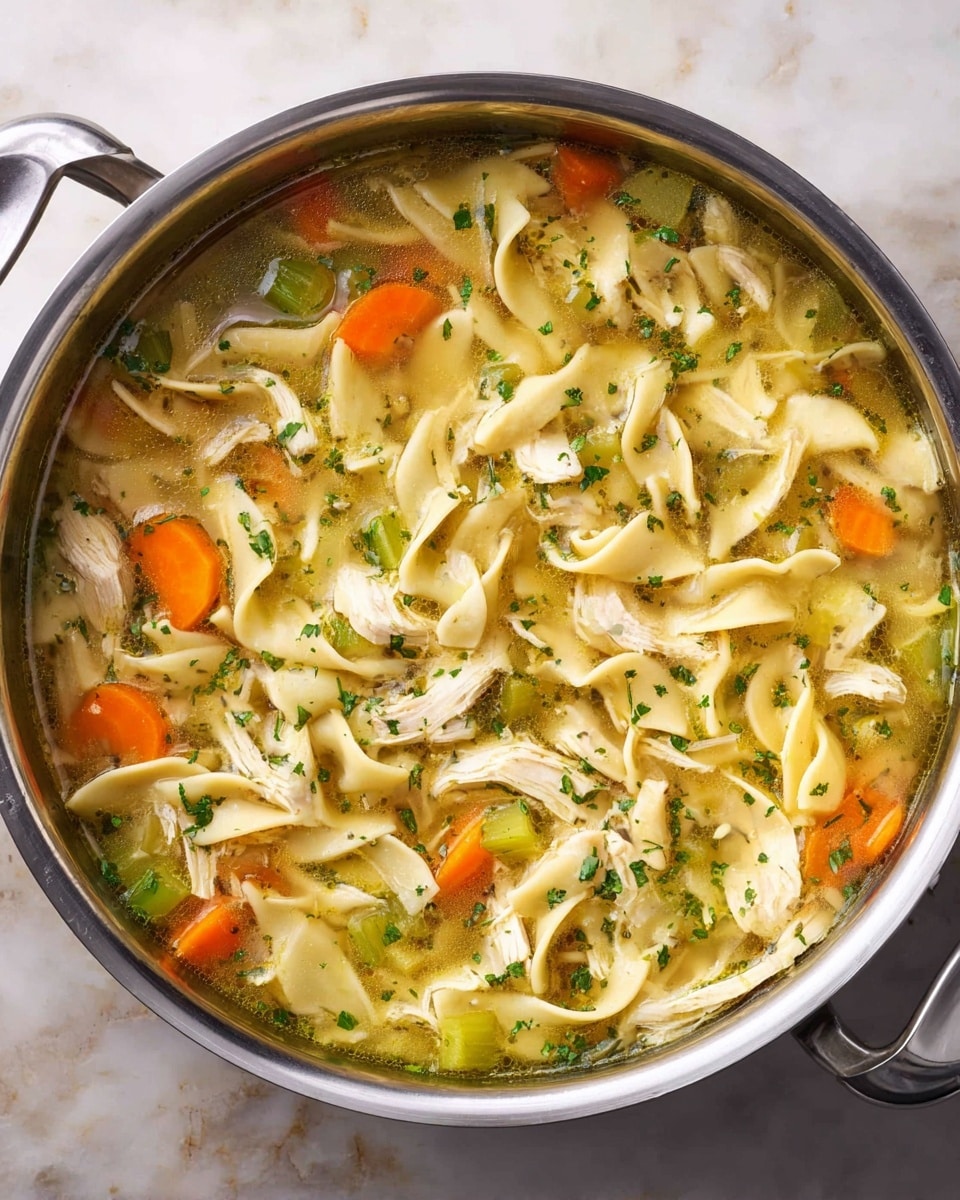 A white bowl filled with chicken noodle soup sits on a white marbled surface. The soup has clear broth with visible small pieces of green celery and orange carrot chunks scattered evenly throughout. Twisted, thick egg noodles float on top, mixed with shredded chicken pieces. The soup’s surface shows small sprinkled herbs, adding texture and green flecks. In the background, there are slices of light brown bread and green leafy parsley partly visible. A silver spoon lays next to the bowl. The photo taken with an iphone --ar 4:5 --v 7