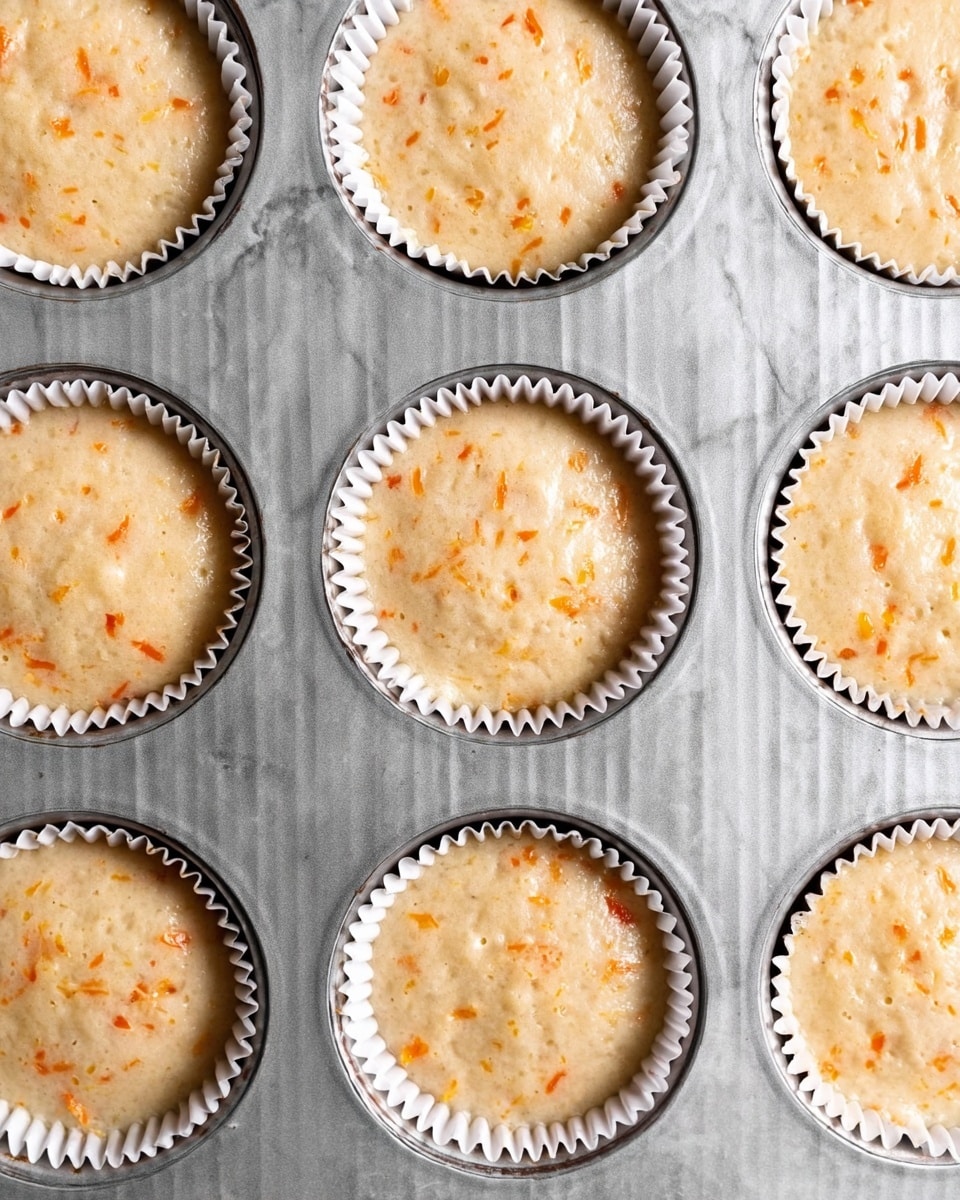 A close-up top view of a metal muffin tray holding twelve white paper liners filled with light beige batter speckled with small orange shreds, each liner evenly filled and smooth on top. The background is a white marbled surface, creating a clean and bright look. The batter is slightly bubbly with a soft, moist texture visible throughout all muffins photo taken with an iphone --ar 4:5 --v 7