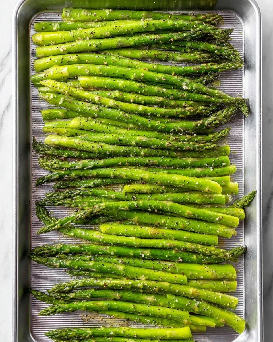 The image shows a silver metal baking tray filled with fresh green asparagus spears. The asparagus is neatly laid out in three rows, covering the tray side to side. Each spear is coated lightly with visible small bits of seasoning, a glistening layer of oil, and specks of black pepper. The asparagus tips are darker green and tightly closed, while the stalks are smooth and bright green. The metal tray has subtle ridges and a slightly shiny surface. The background is a white marbled texture. photo taken with an iphone --ar 4:5 --v 7