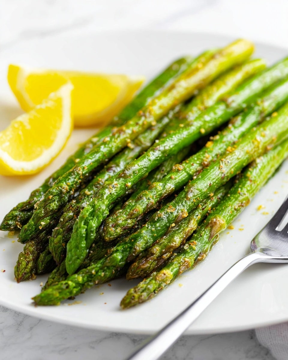 A white plate holds a neat stack of cooked asparagus spears, all bright green with slight char marks showing some crisp roasted texture. The asparagus is arranged in a mostly parallel pile with a small sprinkle of coarse seasoning visible on top. In the background, a lemon wedge with two bright yellow segments sits near the upper left edge of the plate. A silver fork rests on the plate's right side, and the setting is on a white marbled surface. Photo taken with an iphone --ar 4:5 --v 7