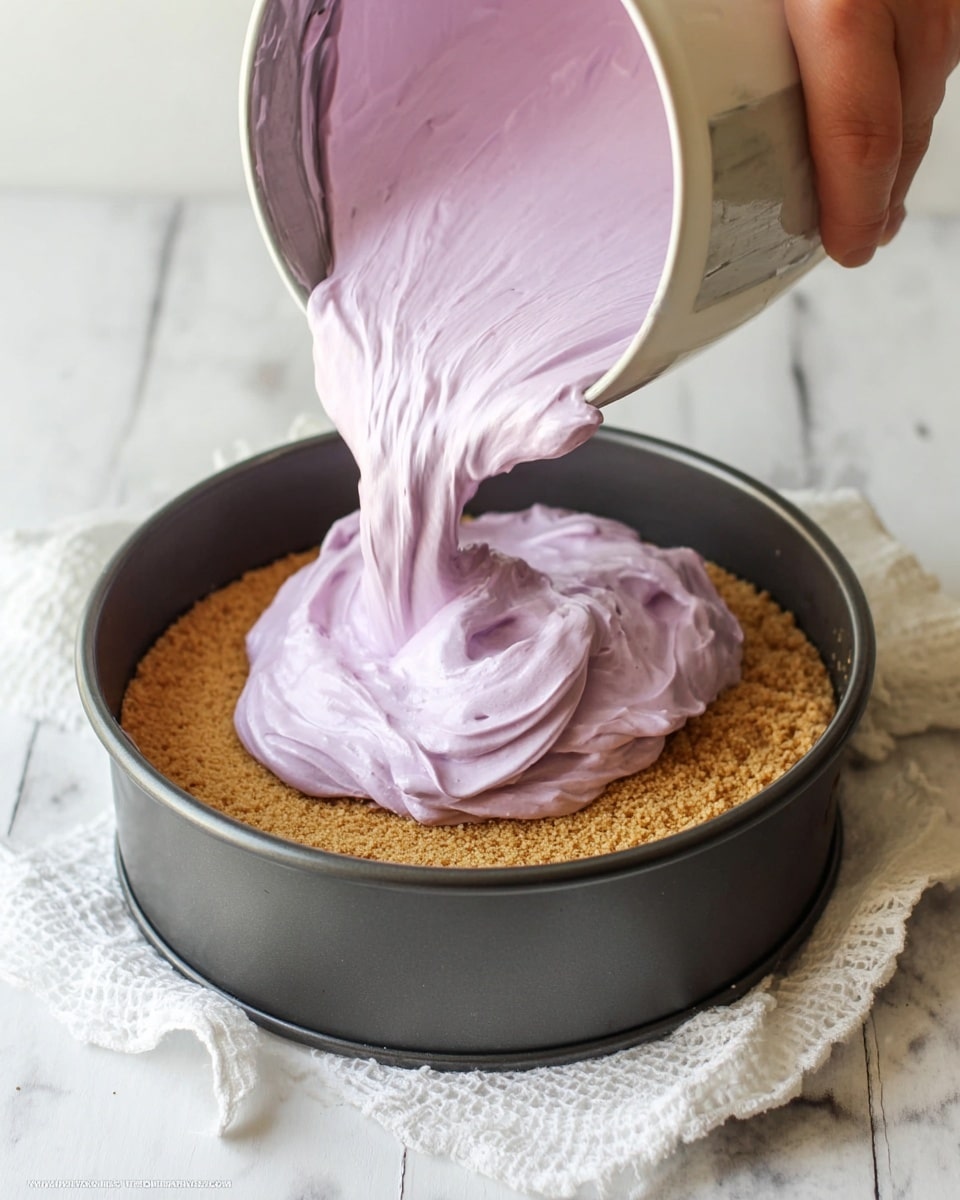 A dark round springform pan holds a single thick layer of light brown crumbly crust at the bottom. Above it, a thick and fluffy light purple creamy mixture is being poured from a shiny metal bowl by a woman's hand holding a spatula, with soft swirls and folds in the creamy layer. The setup rests on a white marbled surface with a white textured cloth nearby. photo taken with an iphone --ar 4:5 --v 7