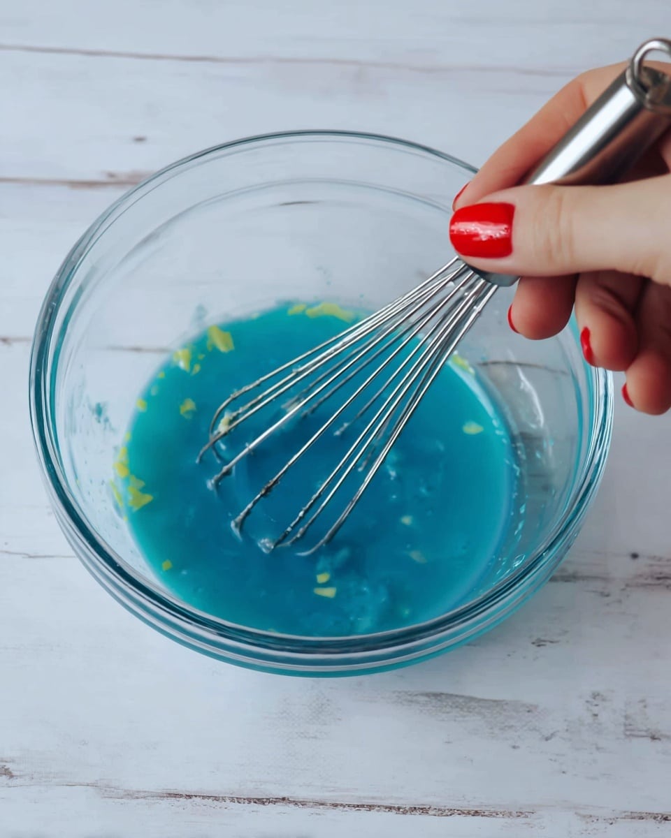 A clear glass bowl with blue liquid inside, being stirred by a small metal whisk held by a woman's hand with red nail polish, the blue liquid is smooth with small yellow bits scattered in it, resting on a white marbled surface photo taken with an iphone --ar 4:5 --v 7