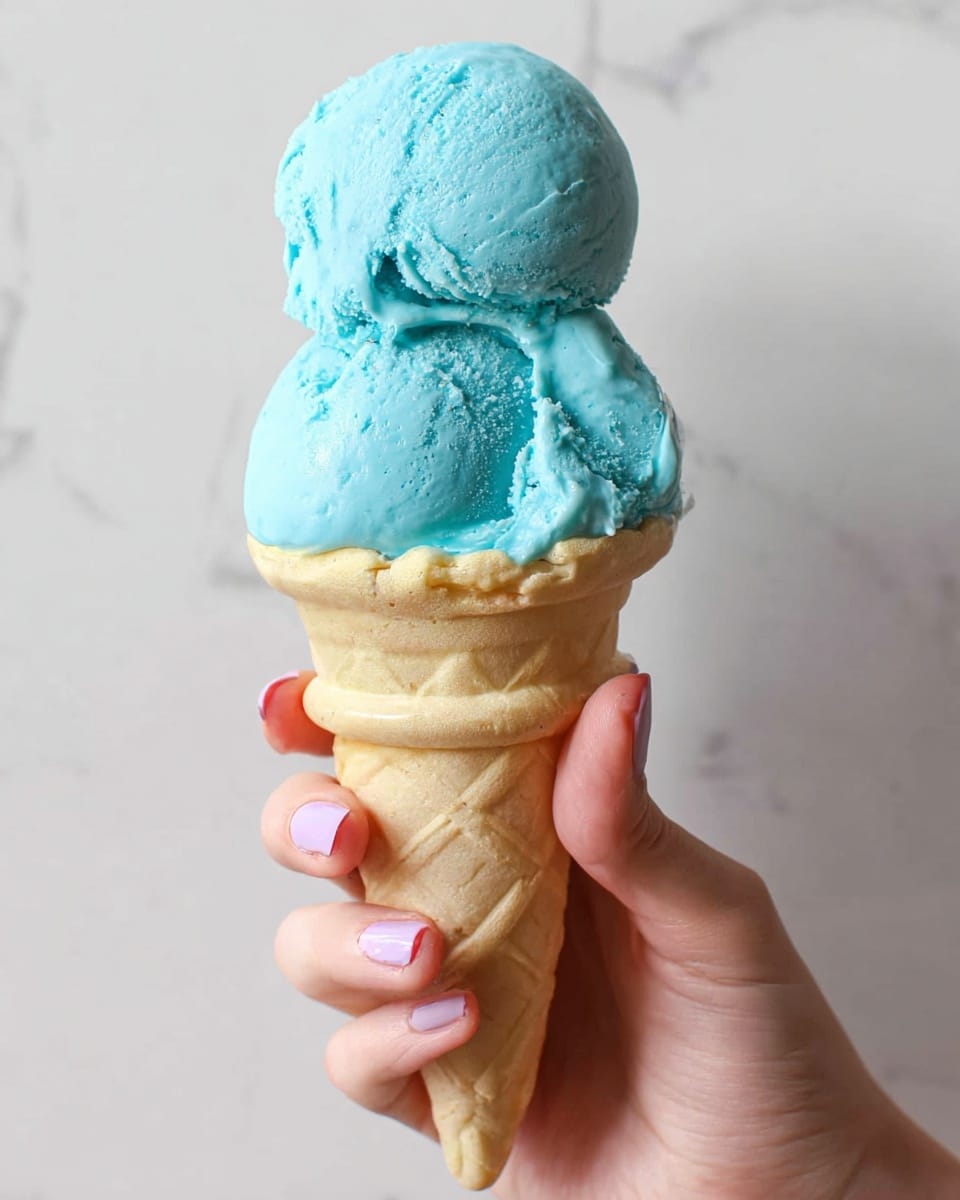 A woman's hand with short nails and light pink polish holds a classic light brown waffle cone with two round scoops of bright blue ice cream stacked on top. The ice cream has a smooth, creamy texture with some soft peaks and gentle swirls, showing a slightly matte finish. The background is a clean white marbled texture, which makes the colors of the ice cream and cone stand out clearly. photo taken with an iphone --ar 4:5 --v 7