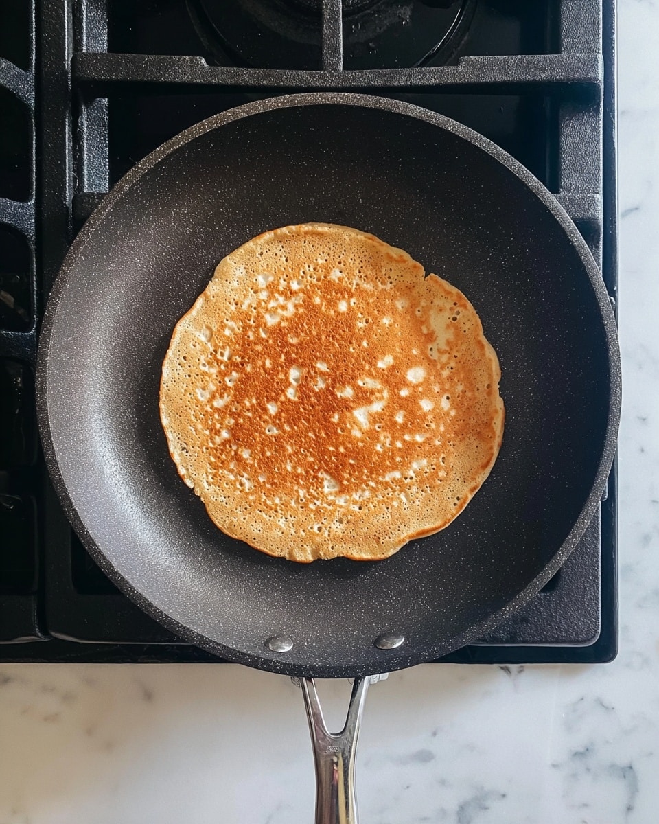 A single round pancake with a light golden brown color and small bubbles on its surface is cooking in a dark gray non-stick frying pan. The pancake is centered in the pan, which rests on a black gas stove grate. The pancake's texture looks fluffy and slightly uneven with a smooth edge. The background has a white marbled texture. photo taken with an iphone --ar 4:5 --v 7