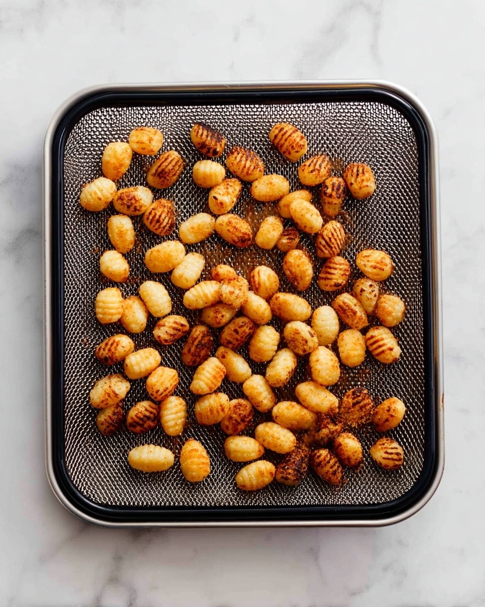 This image shows a single layer of small, golden-brown gnocchi scattered on a square black metal mesh tray. Each gnocchi piece is oval-shaped with some having ridges, and they are toasted to different shades of brown with some darker spots, giving a crispy texture. The tray sits on a white marbled surface, adding cleanliness and brightness to the scene. photo taken with an iphone --ar 4:5 --v 7
