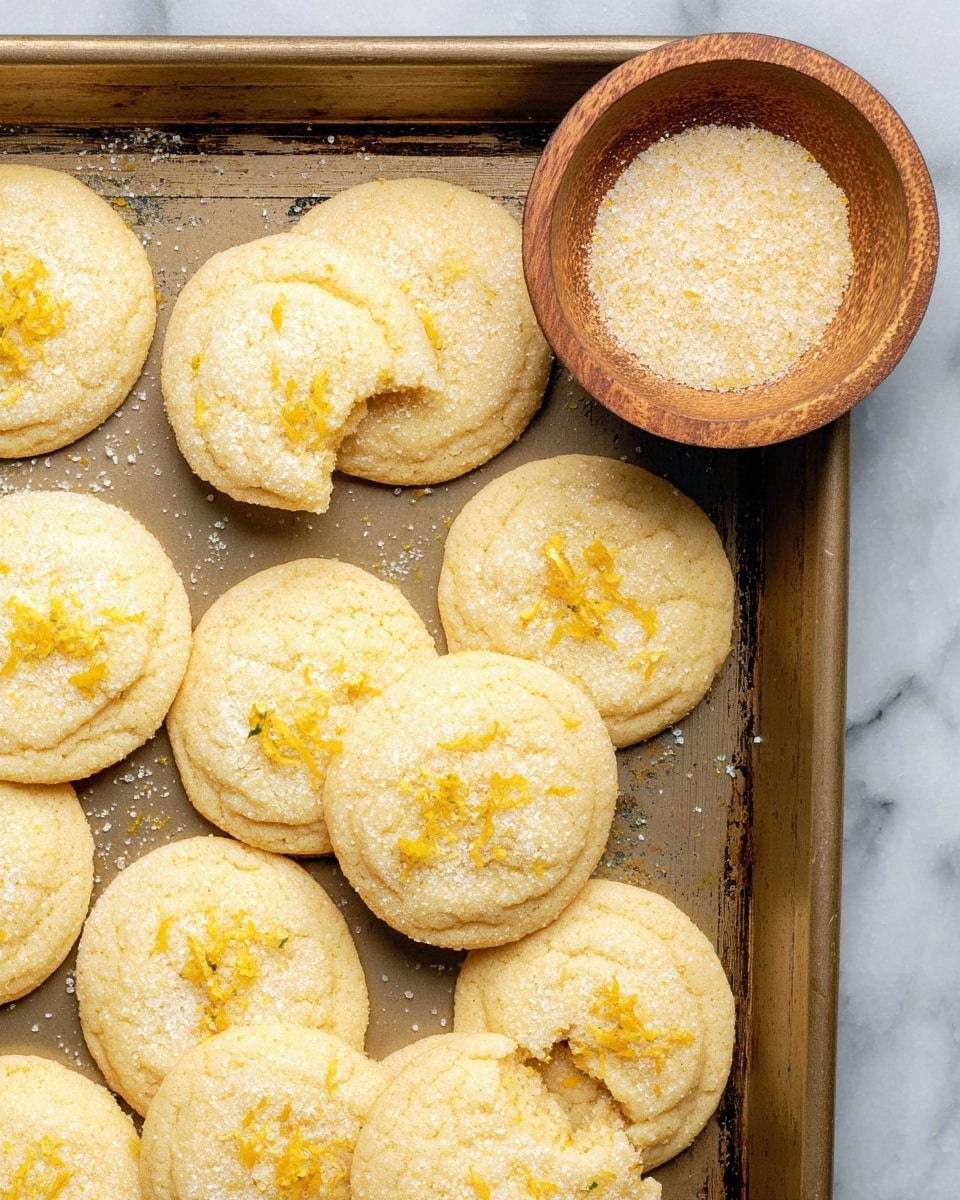 The image shows a metal baking tray filled with round lemon sugar cookies, each cookie having a light golden color with sparkling sugar coating and small bits of lemon zest scattered on top. One cookie near the center has a bite taken out of it, revealing its soft texture inside. Next to the cookies on the top right side of the tray is a small wooden bowl filled with a mix of granulated sugar and lemon zest. The tray is set against a white marbled surface. photo taken with an iphone --ar 4:5 --v 7