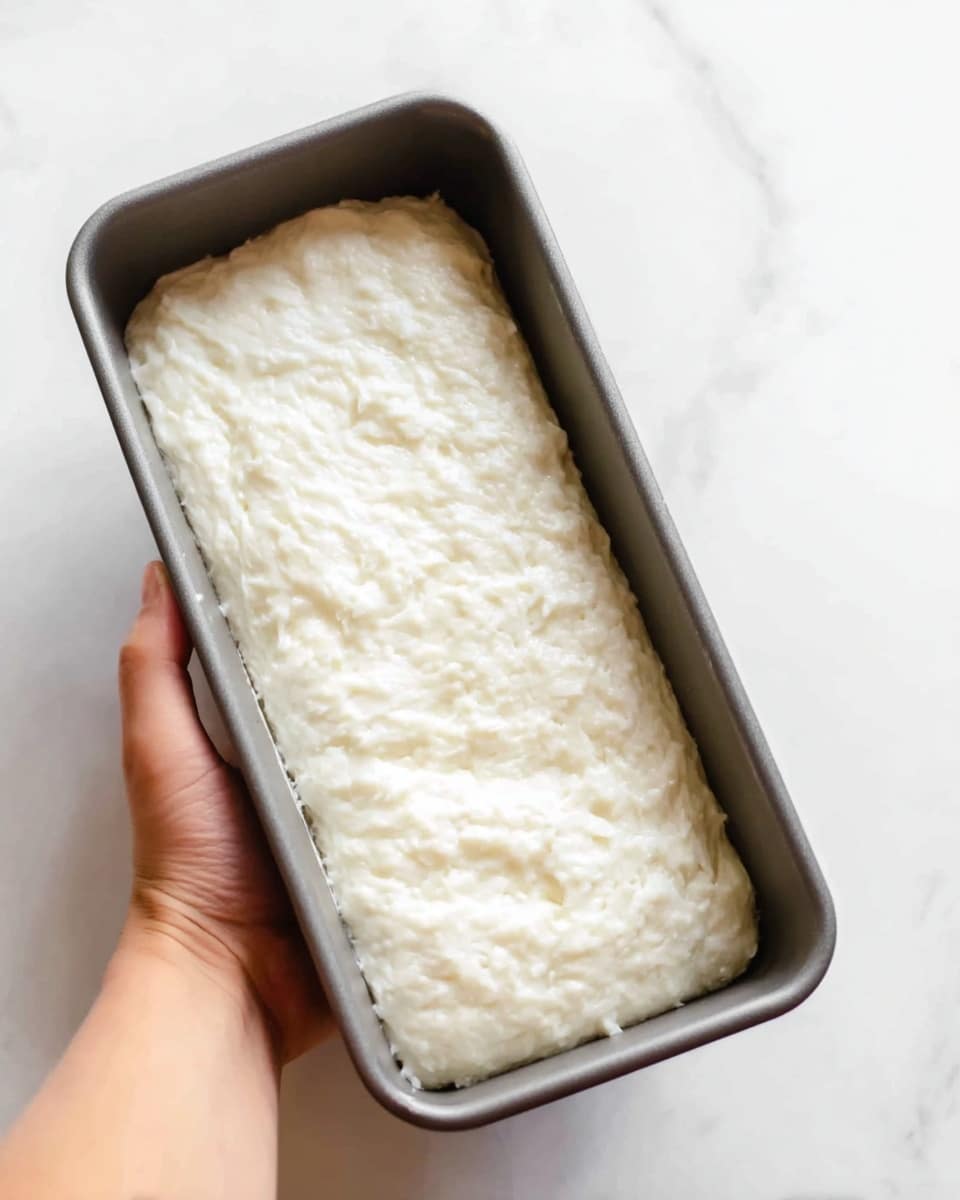 A close-up image showing a woman's hand holding a gray rectangular baking pan with a thick, white dough inside. The dough fills most of the pan, with a soft texture that looks slightly bumpy and stretched, having an even layer covering the entire bottom and sides of the pan. The background is a white marbled surface. Photo taken with an iphone --ar 4:5 --v 7