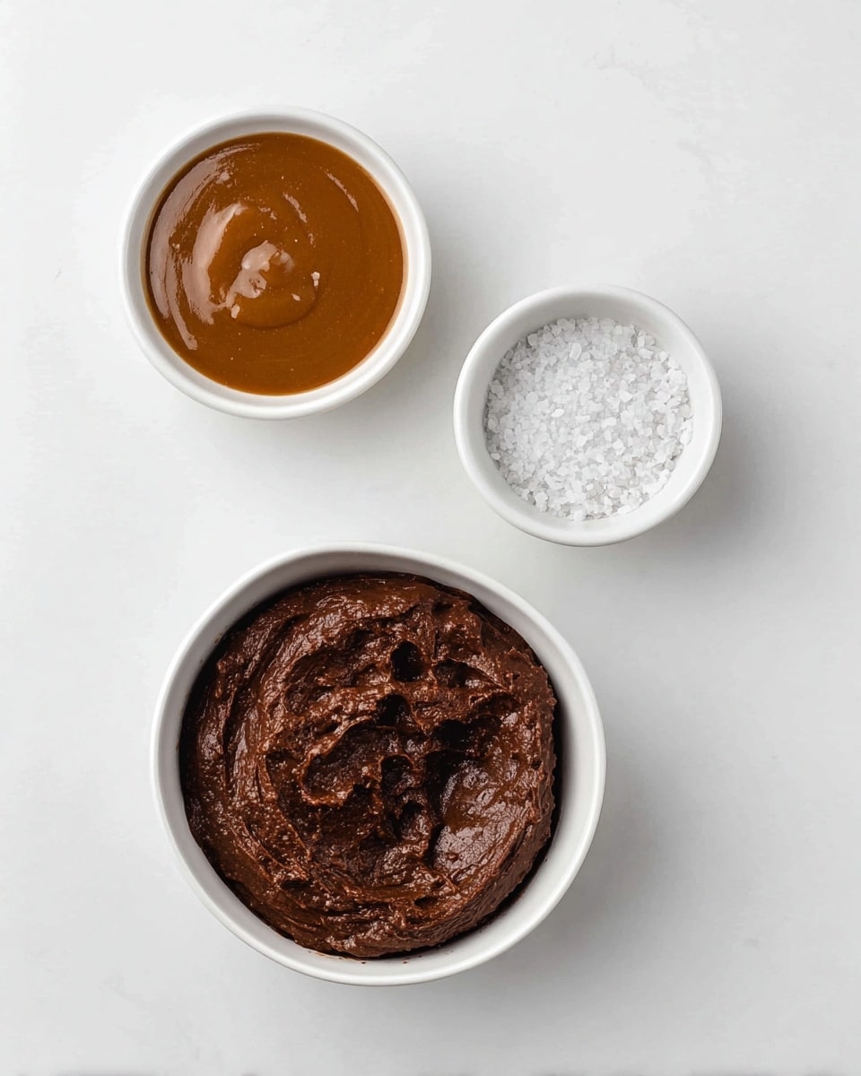 The image shows three white bowls placed on a white marbled surface. The largest bowl, positioned at the bottom, contains thick, dark brown chocolate batter with a rough, creamy texture. Above it, slightly to the left, there is a medium bowl filled with smooth brown caramel sauce. To the right of the caramel, a smaller white bowl holds coarse grains of white salt, creating a contrast with the dark bowls. The items are arranged in a simple and clean layout with soft lighting. photo taken with an iphone --ar 4:5 --v 7
