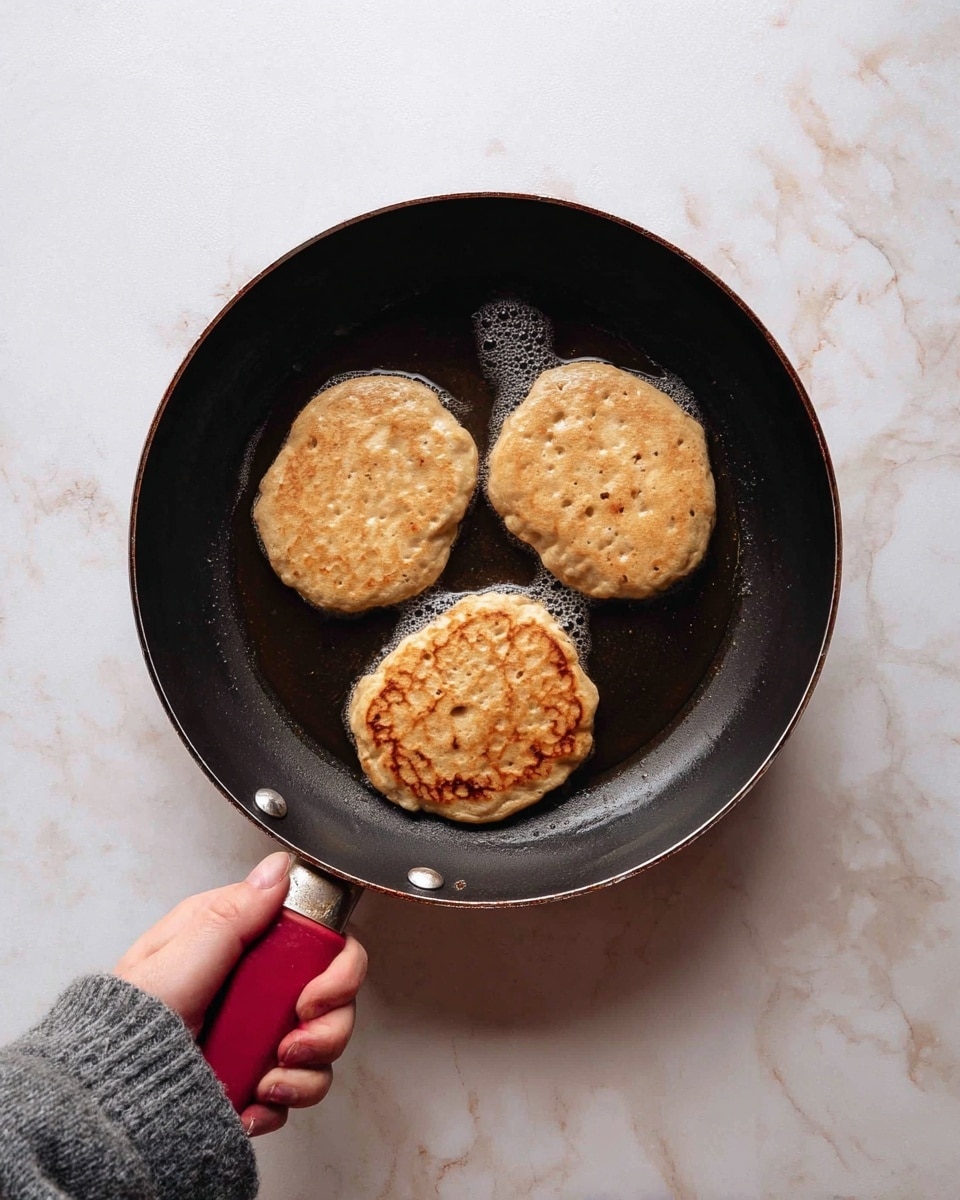 A black frying pan held by a woman's hand in a grey sleeve contains three thick, round, bubbling pancakes cooking in hot oil. Each pancake is light brown with a slightly uneven texture and small holes on the surface, showing they are cooking. The pan has a dark red handle and is placed over a white marbled surface background. photo taken with an iphone --ar 4:5 --v 7
