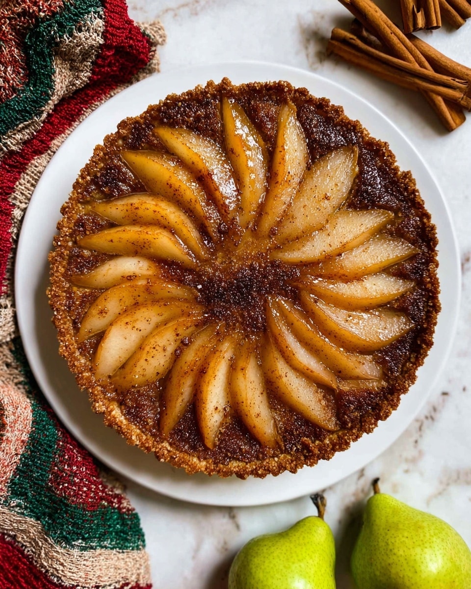 A round tart with two layers sits on a white plate on a white marbled surface. The bottom layer is a crumbly brown crust, topped with a layer of sliced golden-brown pears arranged in a circular pattern starting from the outer edge and moving inward. The pear slices have a shiny glaze with specks of dark cinnamon or spice sprinkled over them. The center is filled with a slightly darker spiced mixture that blends into the pear slices. Near the plate, two whole cinnamon sticks lay on the surface along with a colorful red, green, and cream woven cloth and two fresh green pears. Photo taken with an iphone --ar 4:5 --v 7