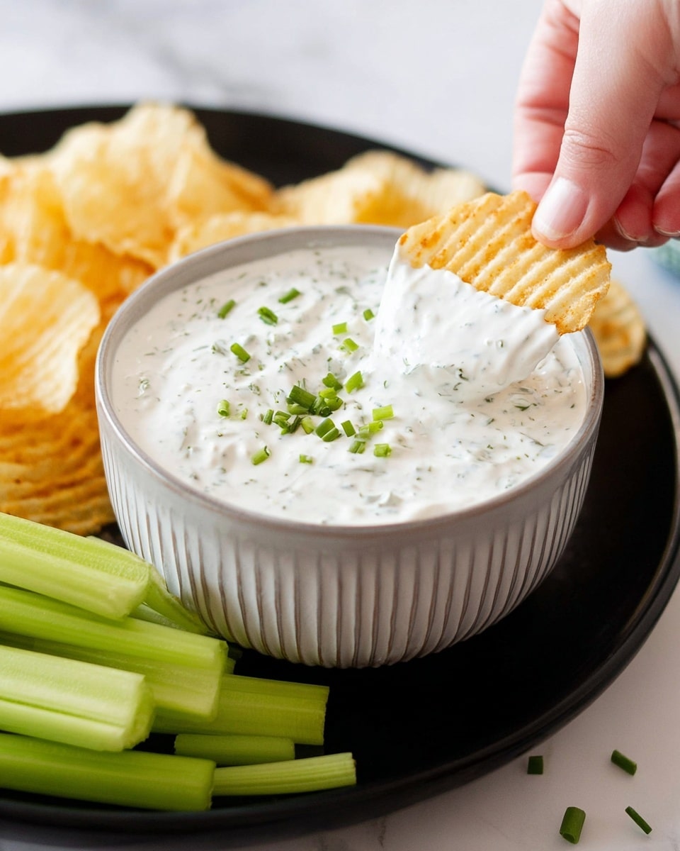 A white ridged bowl filled with creamy white ranch dip speckled with herbs and topped with small green chive pieces sits on a black plate. On the plate around the bowl are light golden wavy potato chips and fresh celery sticks with light and slightly darker green colors. A woman's hand is dipping a potato chip into the ranch dip. The whole scene is placed on a white marbled surface. photo taken with an iphone --ar 4:5 --v 7