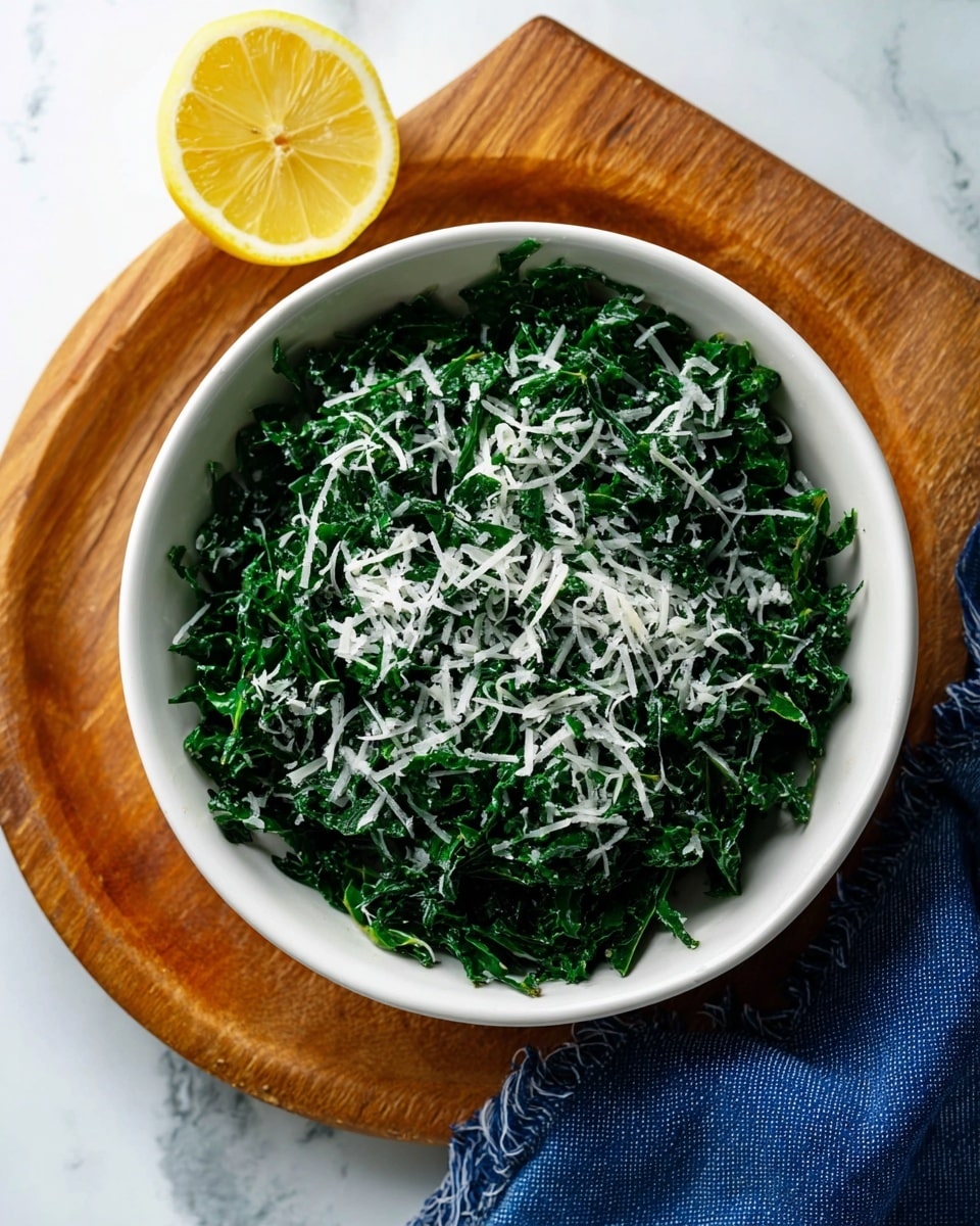 A white scalloped bowl filled with finely chopped dark green kale, lightly dressed and topped with small pieces of shredded white cheese scattered evenly on top. A vintage silver fork with an ornamental handle is placed inside the bowl on the right side, resting on the kale. The bowl is set on a white marbled surface next to a folded blue textured cloth, with a yellow lemon half partially visible on the lower left side. Photo taken with an iphone --ar 4:5 --v 7